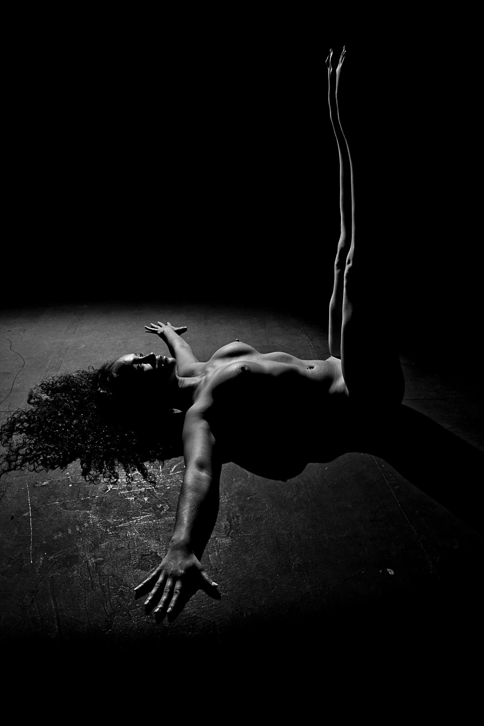 Mujer desnuda tumbada de espaldas sobre un suelo oscuro, con el brazo abierto y las piernas elevadas en vertical, iluminada por un foco.