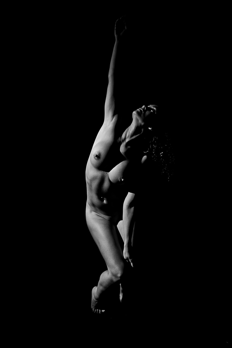 Figura desnuda con el cuerpo arqueado y un brazo estirado hacia arriba, en luz lateral sobre fondo negro.