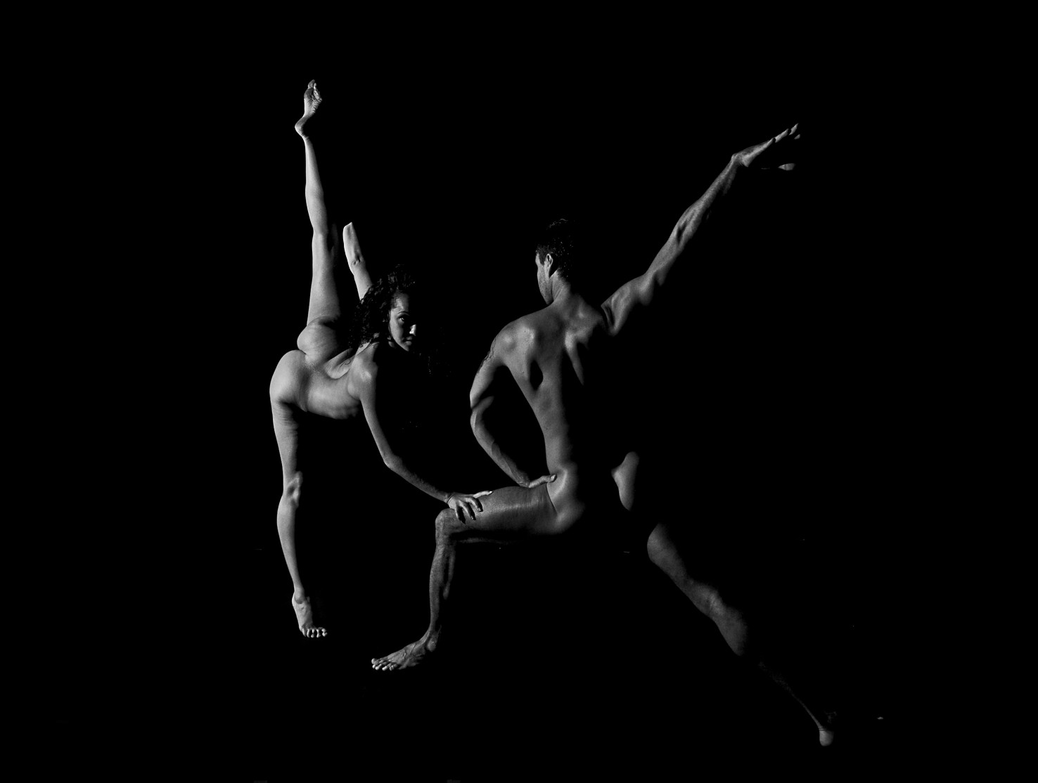 Una bailarina y un bailarín desnudos en movimiento: ella con una pierna extendida hacia arriba, él con un brazo abierto, sobre fondo negro.