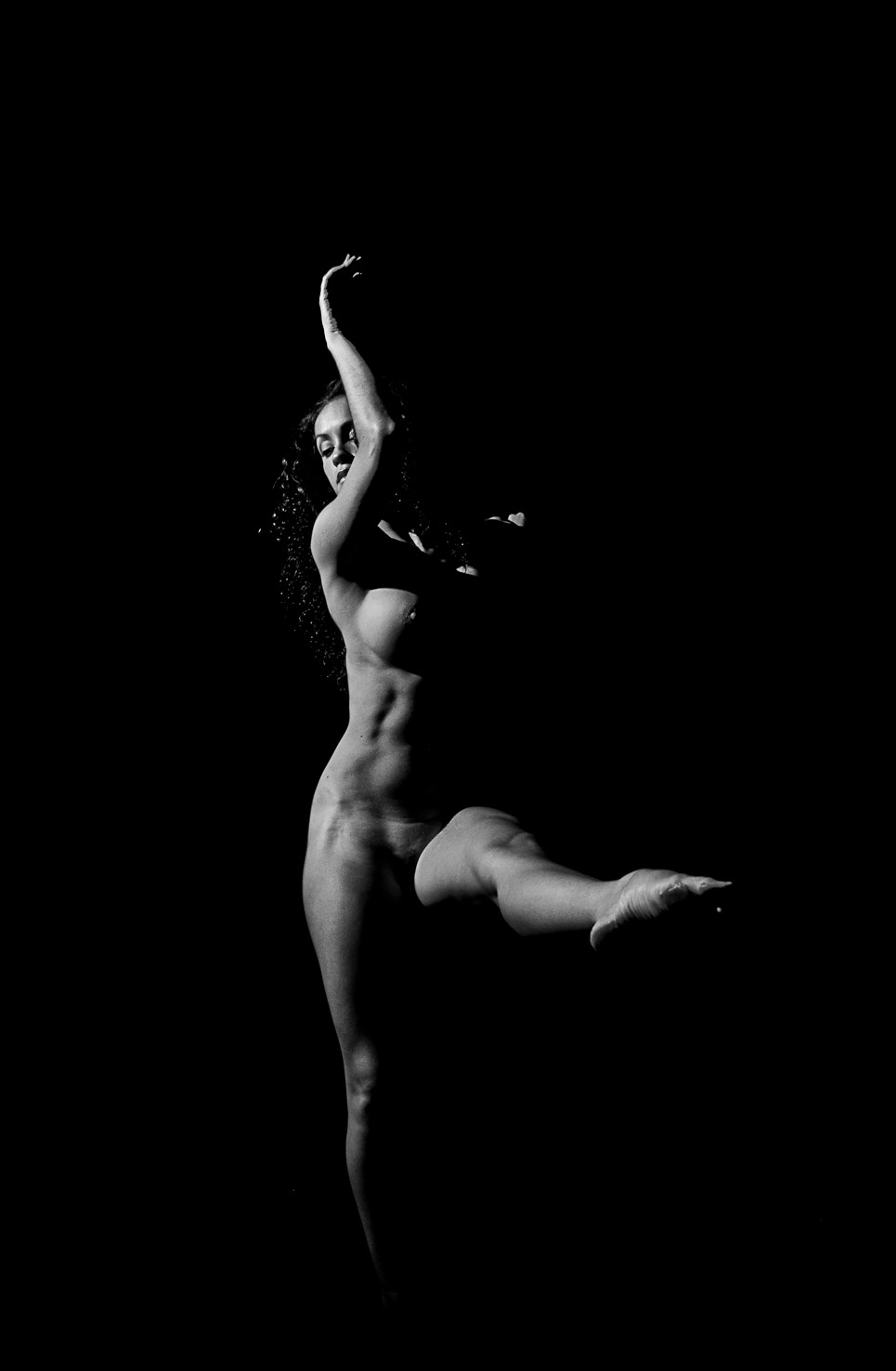 Bailarina desnuda de perfil, con un brazo elevado y una pierna levantada hacia atrás, sobre fondo negro.