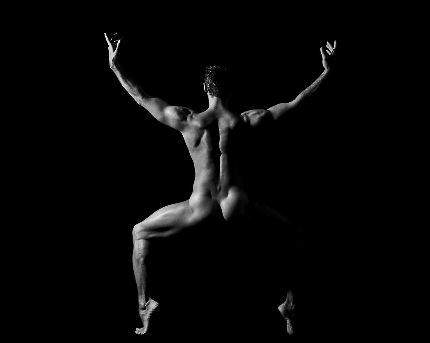 Bailarín desnudo visto de espaldas, con los brazos en alto y las piernas abiertas, sobre fondo negro.