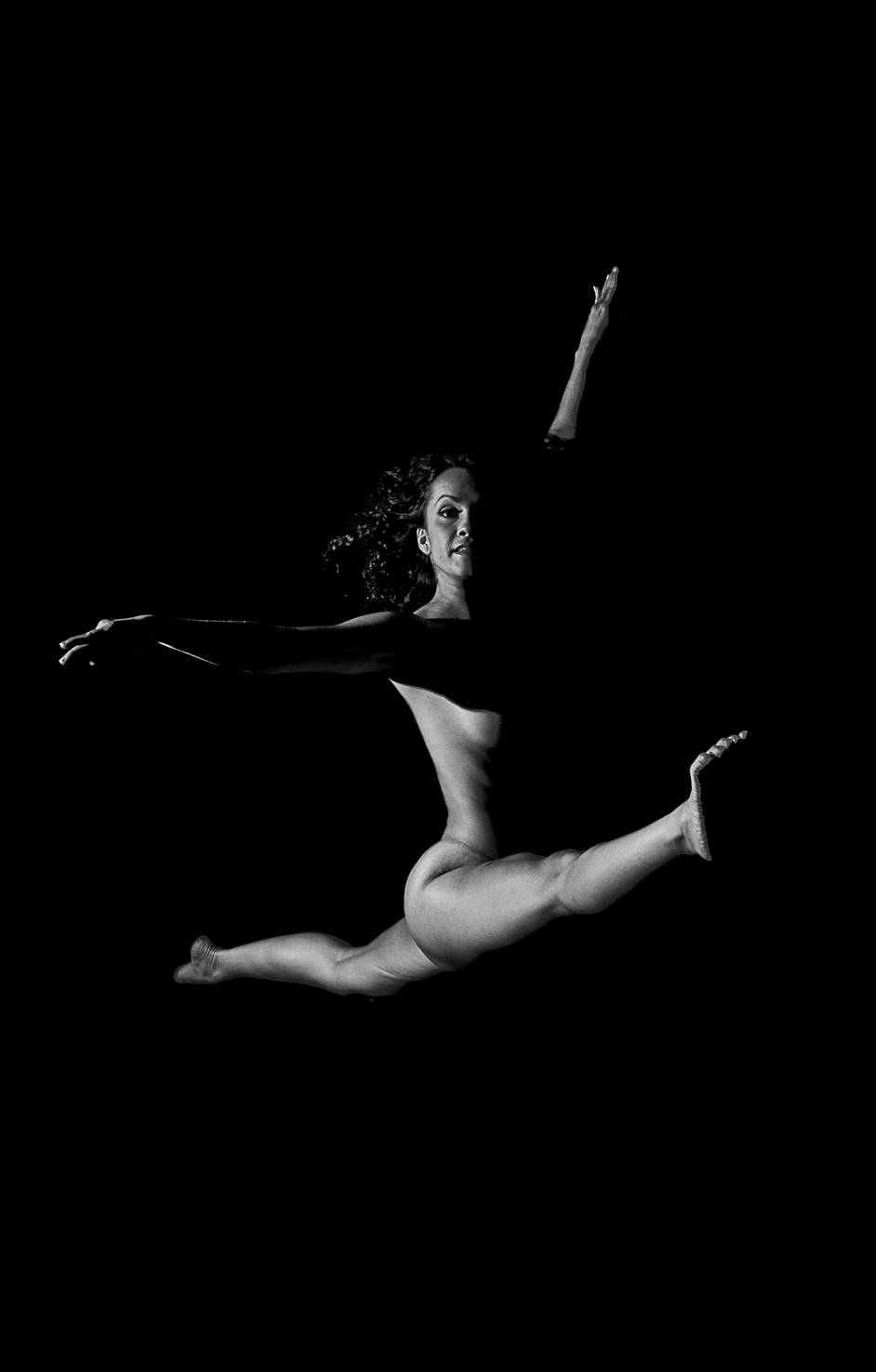 Bailarina desnuda en pleno salto, con las piernas abiertas y un brazo extendido, sobre fondo negro.