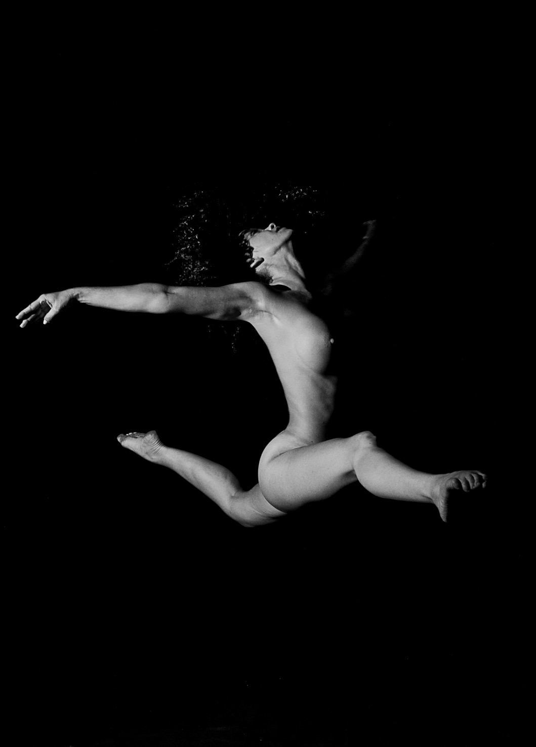 Bailarina desnuda en salto con las piernas abiertas y un brazo estirado hacia un lado, sobre fondo negro.