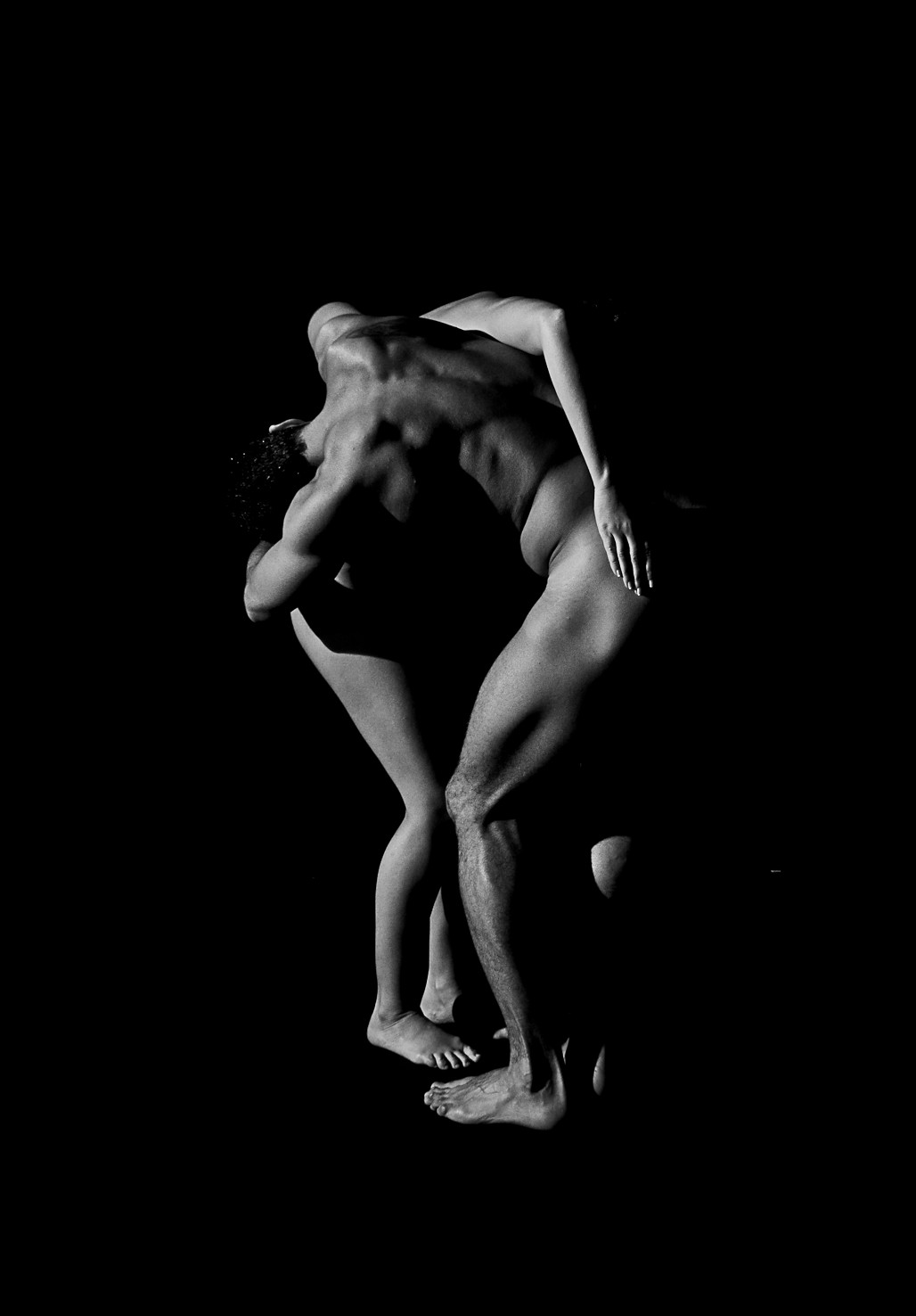 Dos figuras desnudas abrazadas e inclinadas hacia delante, sobre fondo negro.
