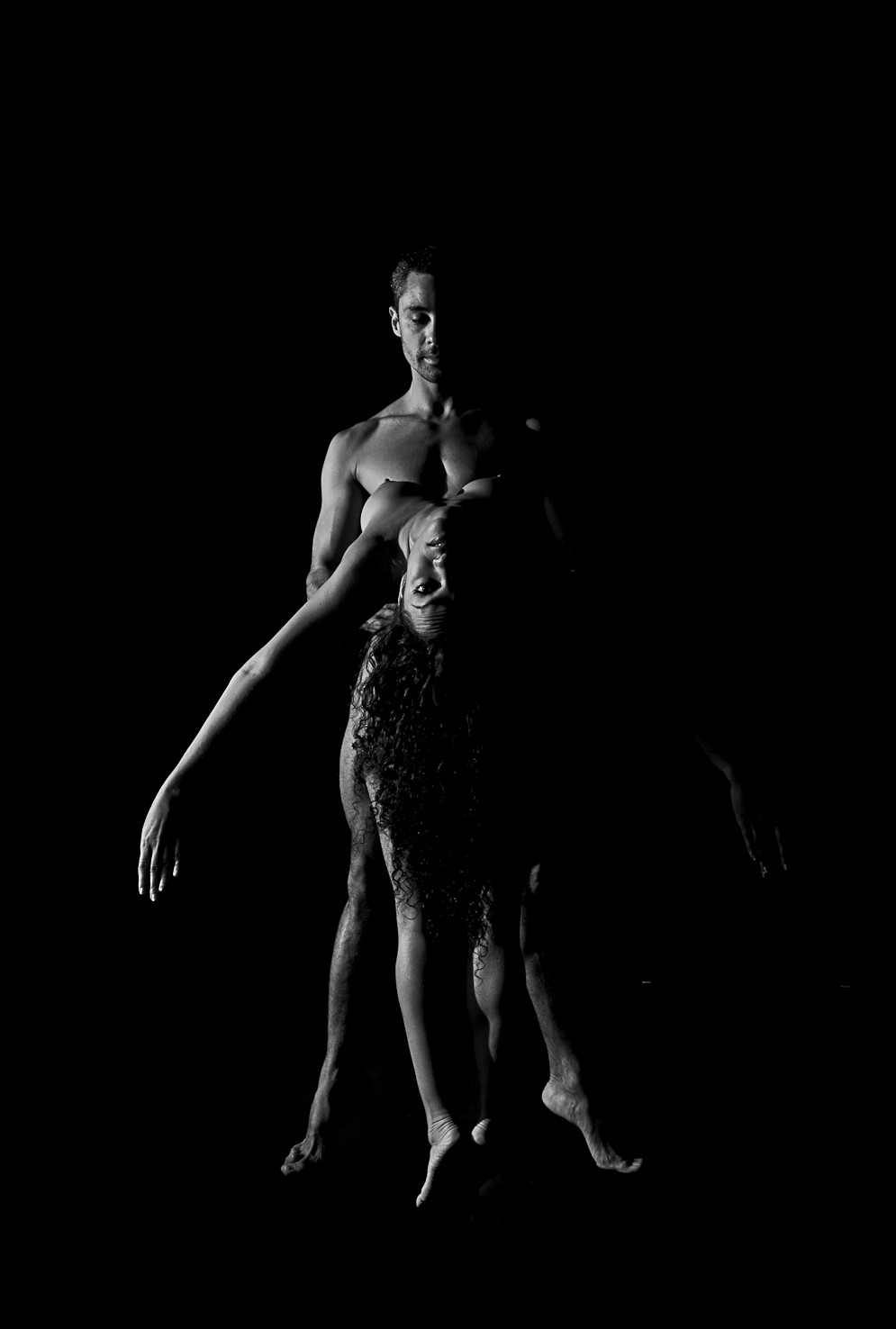 Un hombre y una mujer desnudos de pie, ella delante de él, con un brazo extendido hacia un lado, sobre fondo negro.