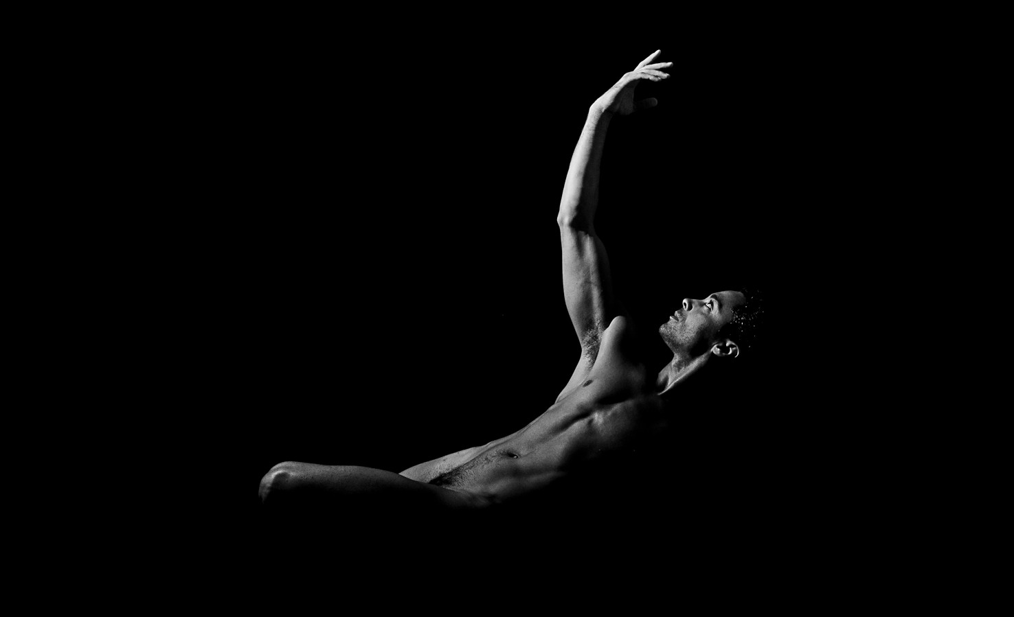 Bailarín desnudo recostado hacia atrás, con un brazo alzado y la cabeza girada, sobre fondo negro.