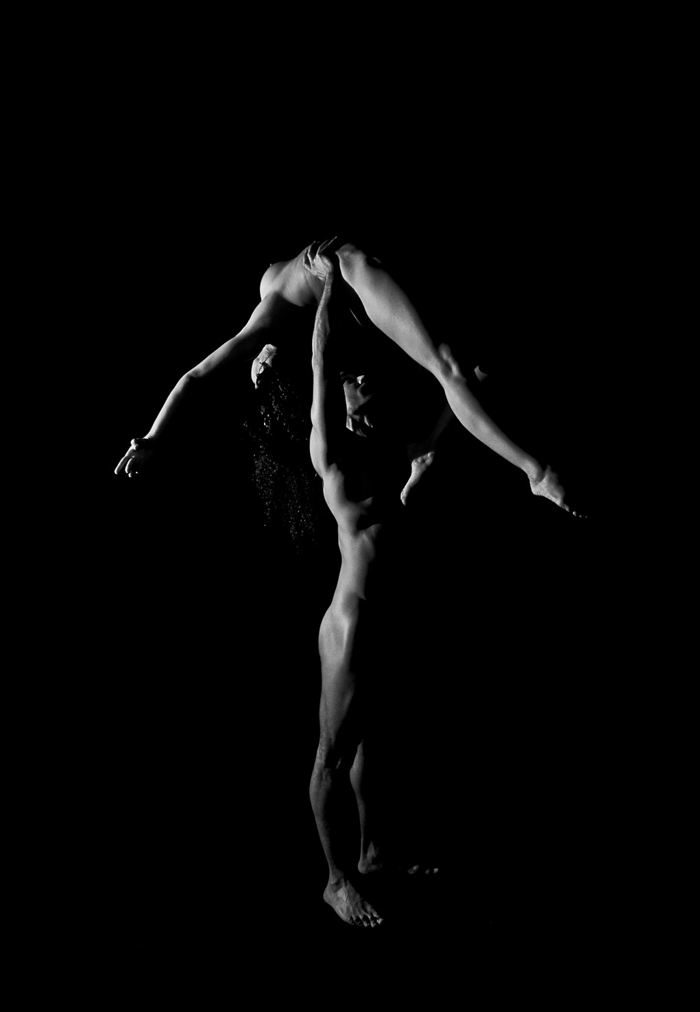 Dos figuras desnudas, una sostenida en alto sobre la otra, con las piernas abiertas y los brazos extendidos, sobre fondo negro.