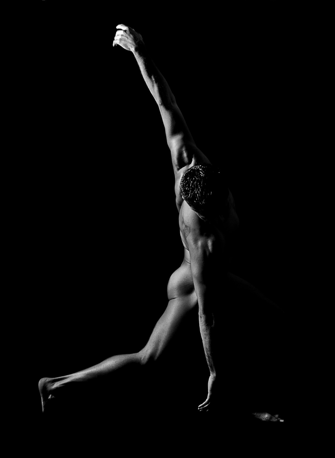 Bailarina desnuda de espaldas dando una zancada larga, con un brazo estirado en vertical, sobre fondo negro.