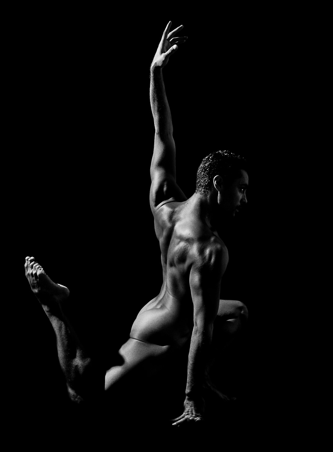 Bailarín desnudo visto de espaldas, agachado sobre una pierna, con un brazo alzado, sobre fondo negro.