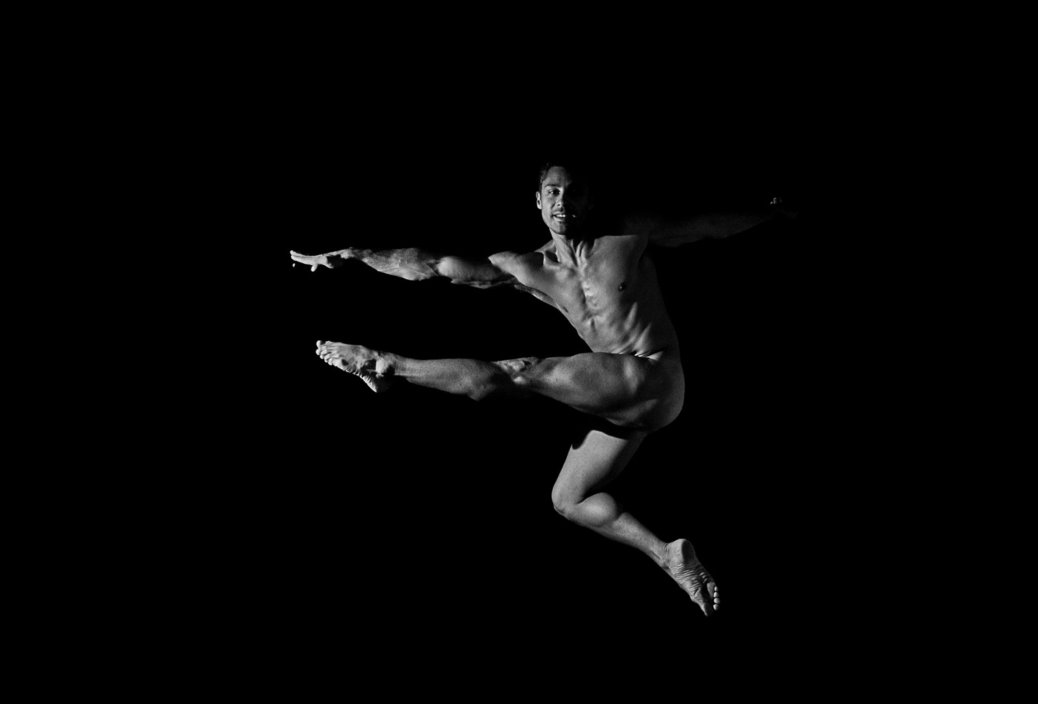 Bailarín desnudo en salto, con las piernas flexionadas, los brazos extendidos y una sonrisa, sobre fondo negro.
