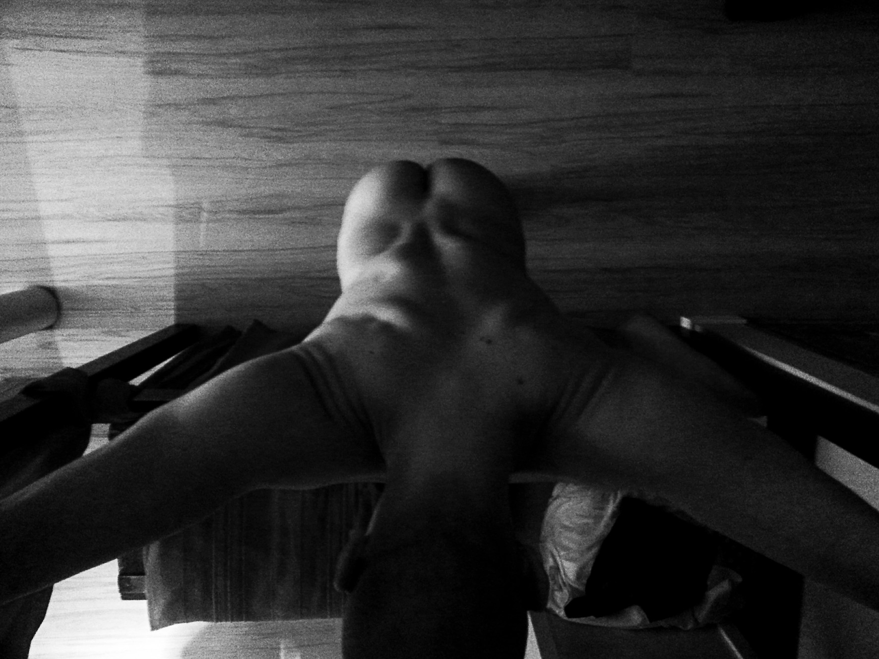 A nude sculpted figure seen from below, arms outstretched, amid shadows and wood.