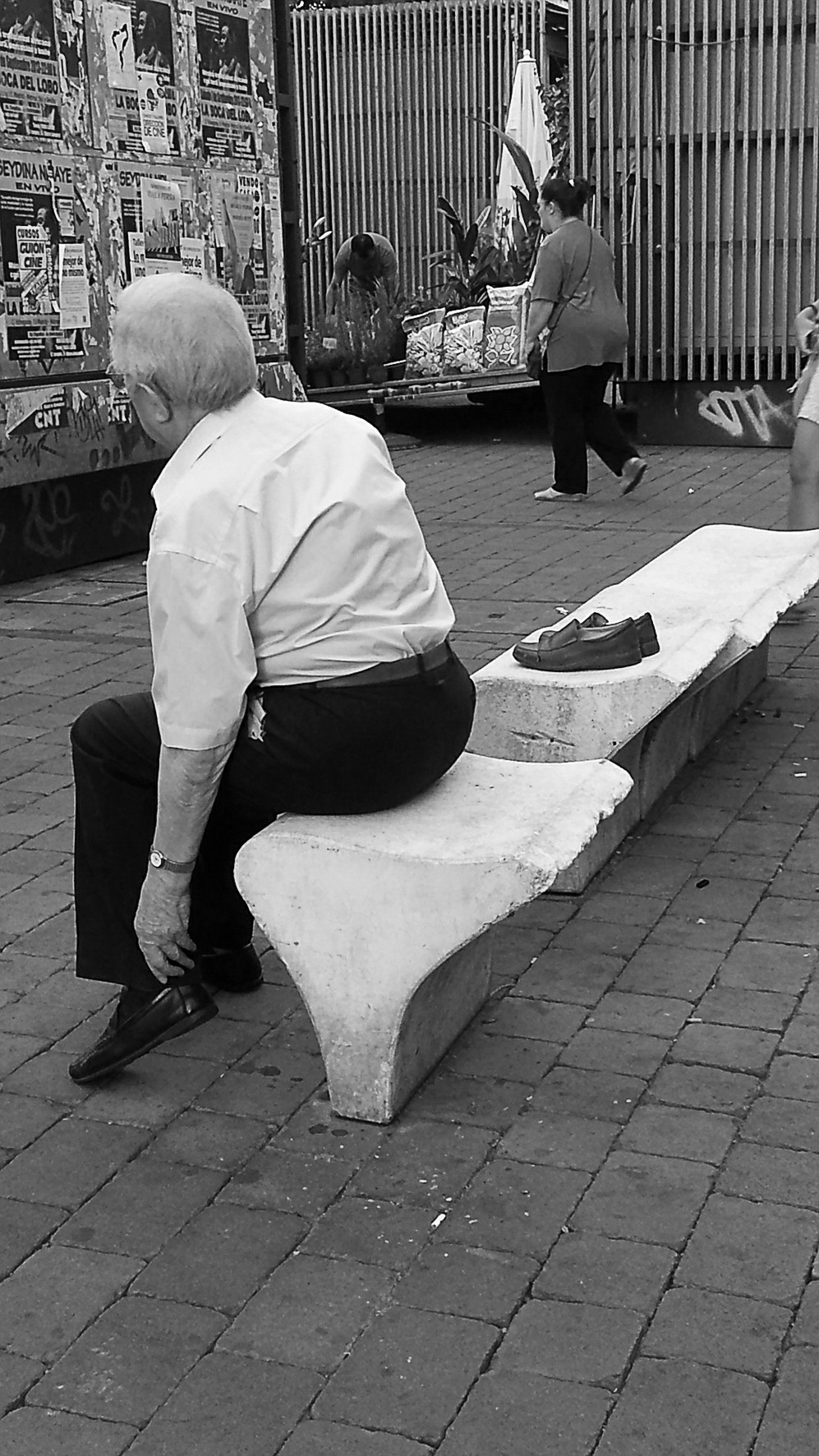 An elderly man seen from behind, seated on a stone bench in a square, posters and a gate behind him.