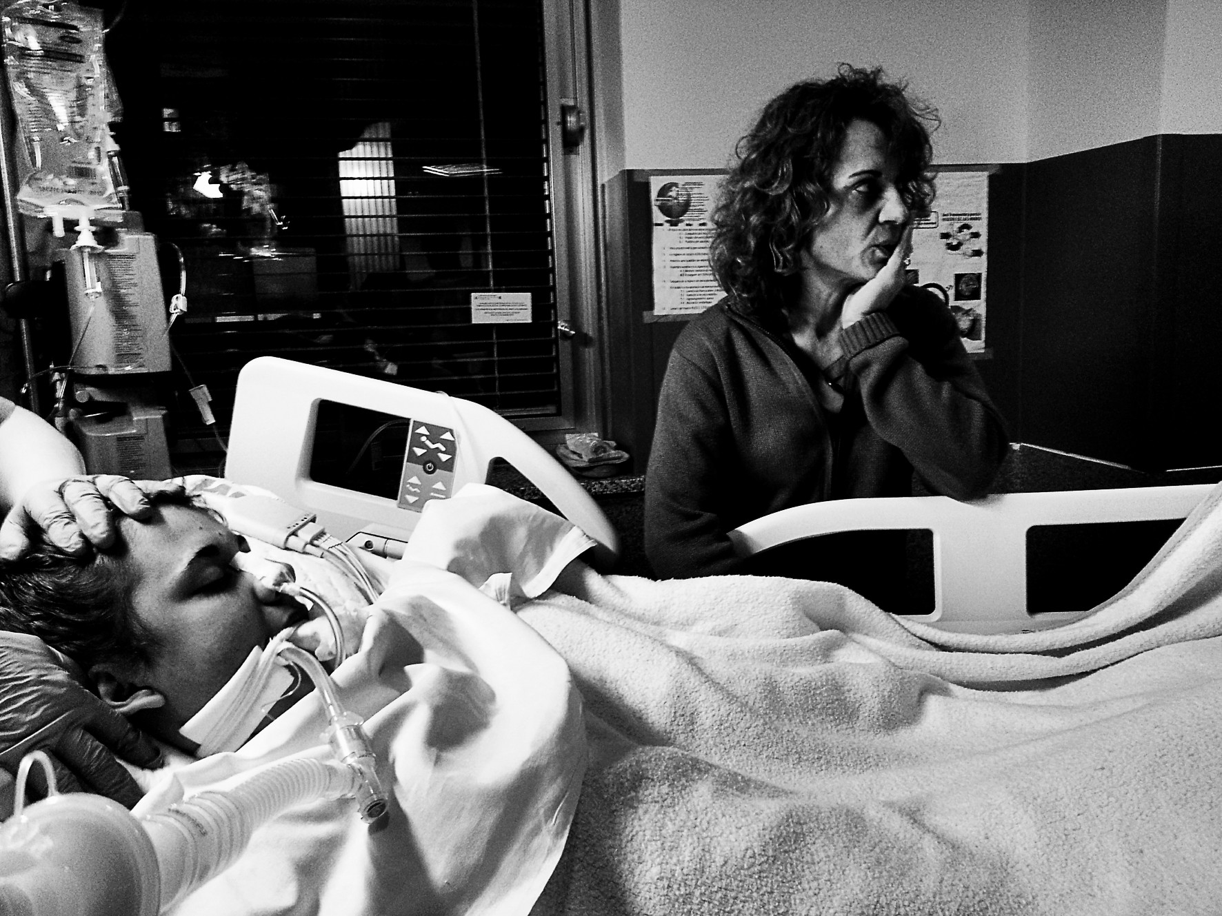 In a hospital room, a man in bed with tubes and monitors; beside him, a woman with her hand to her mouth looks aside.