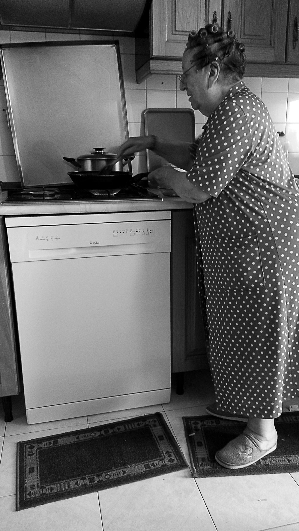 An older woman in a housecoat cooking at a stove in a small kitchen, standing beside a pan.