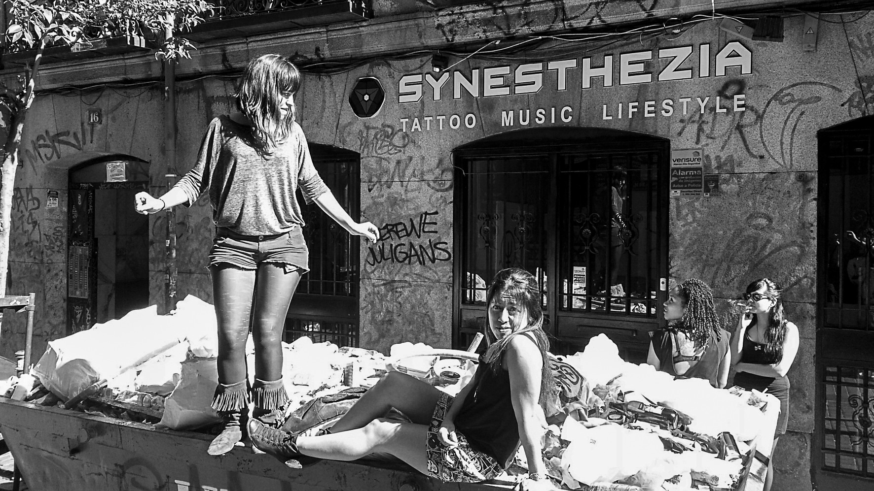 Two women pose on a rubble skip in front of a graffiti-covered shop sign reading "Synesthezia"; two more watch from behind.