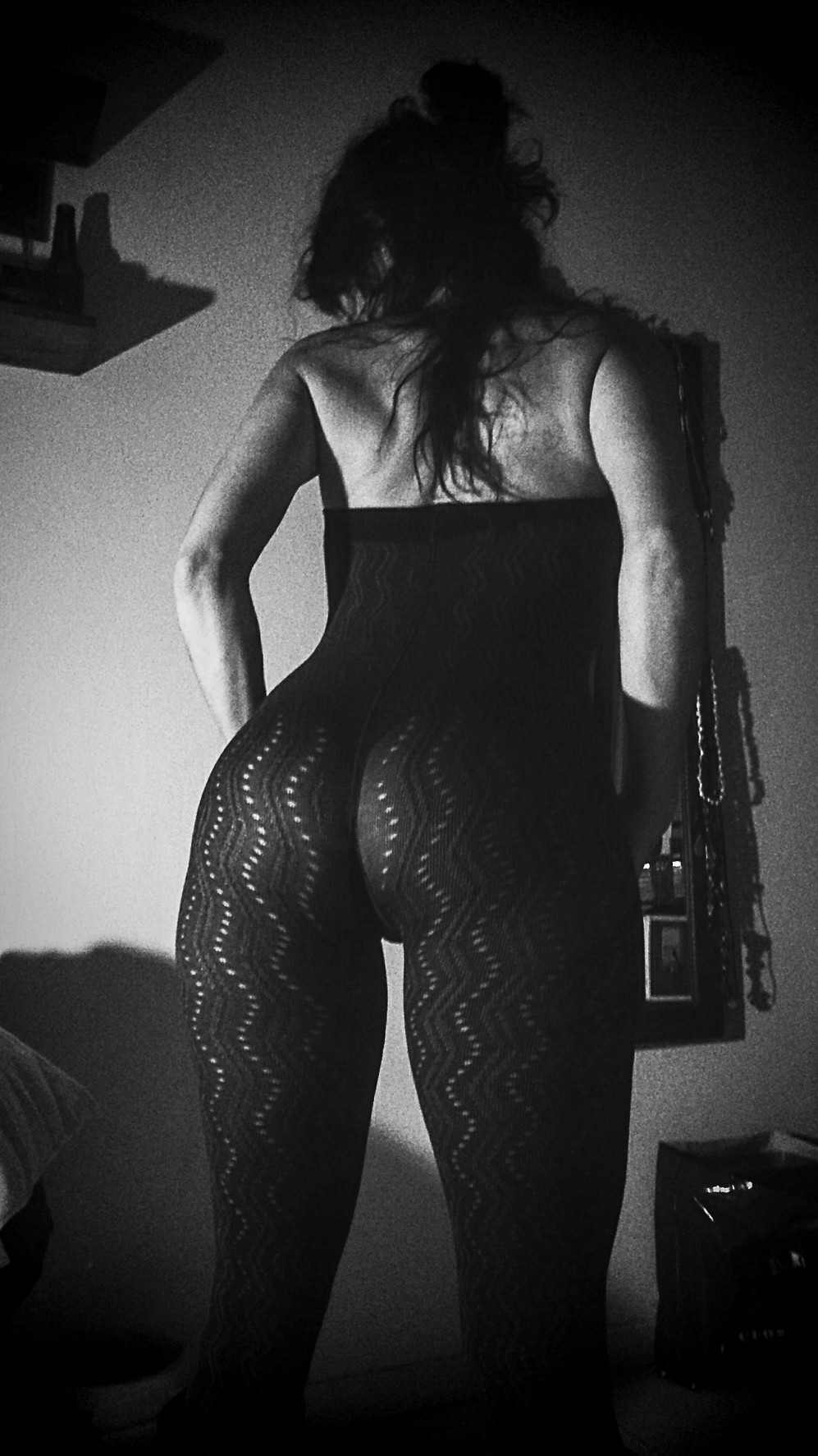 Woman seen from behind in patterned tights and a black bodysuit, an object hanging from her shoulder, in low light.