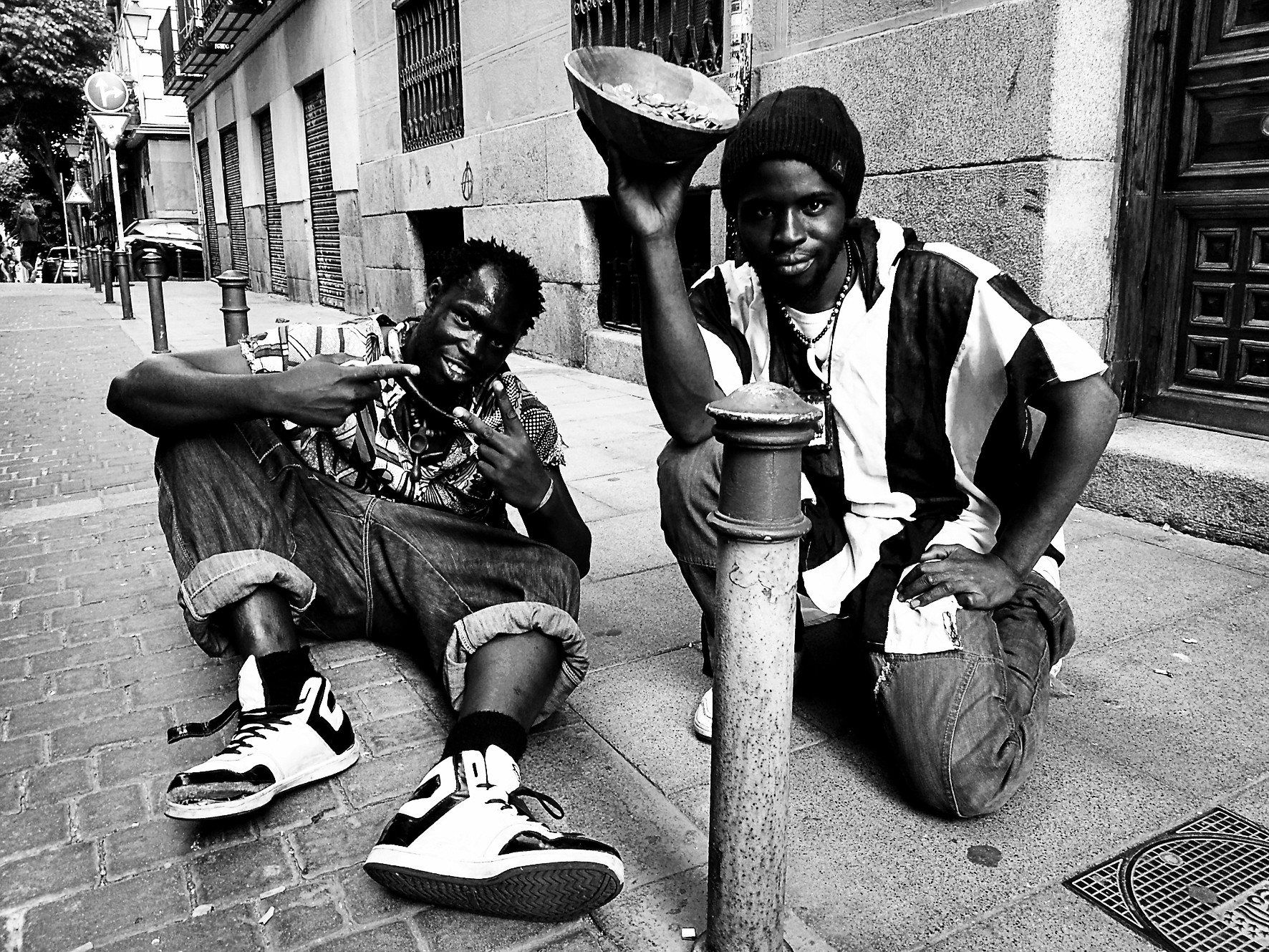 Two young men on a cobbled street: one seated on the ground, the other crouched, holding a bowl aloft.