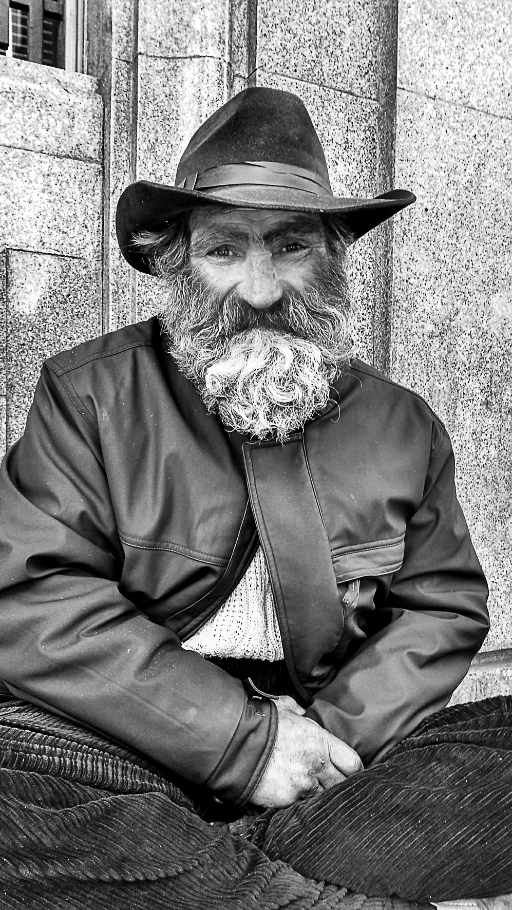 Portrait of an older man in a hat and beard, seated with folded hands beside a wall.