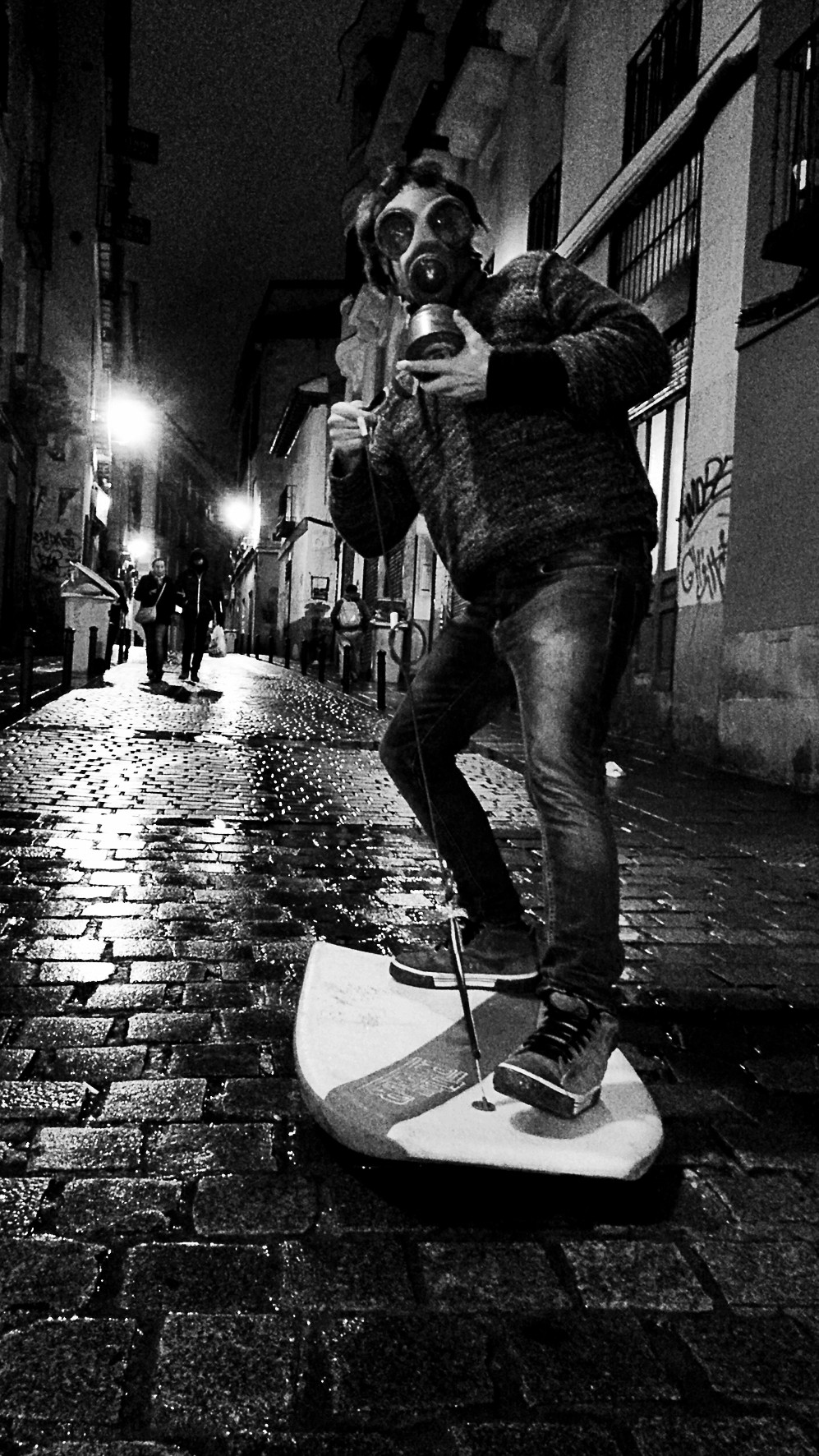 A man in a gas mask balances on a surfboard on a wet cobbled street at night.