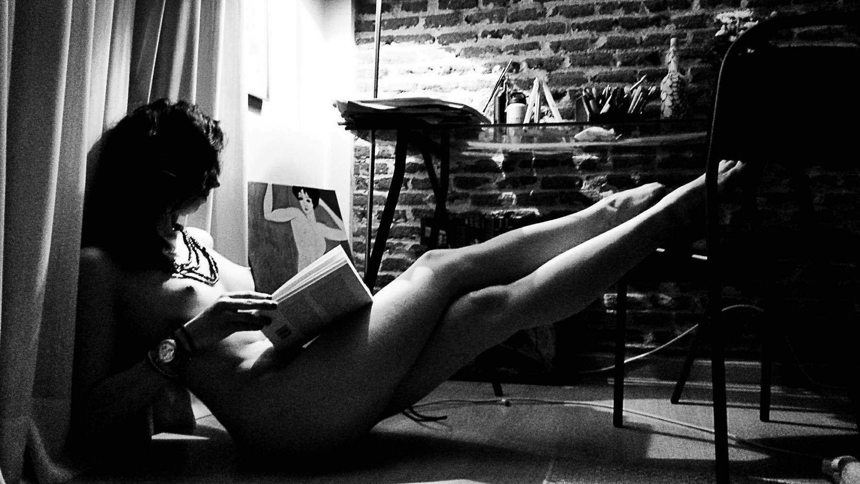 Nude woman reading a book, reclining beside a curtain, her legs resting up high, a painting behind her.