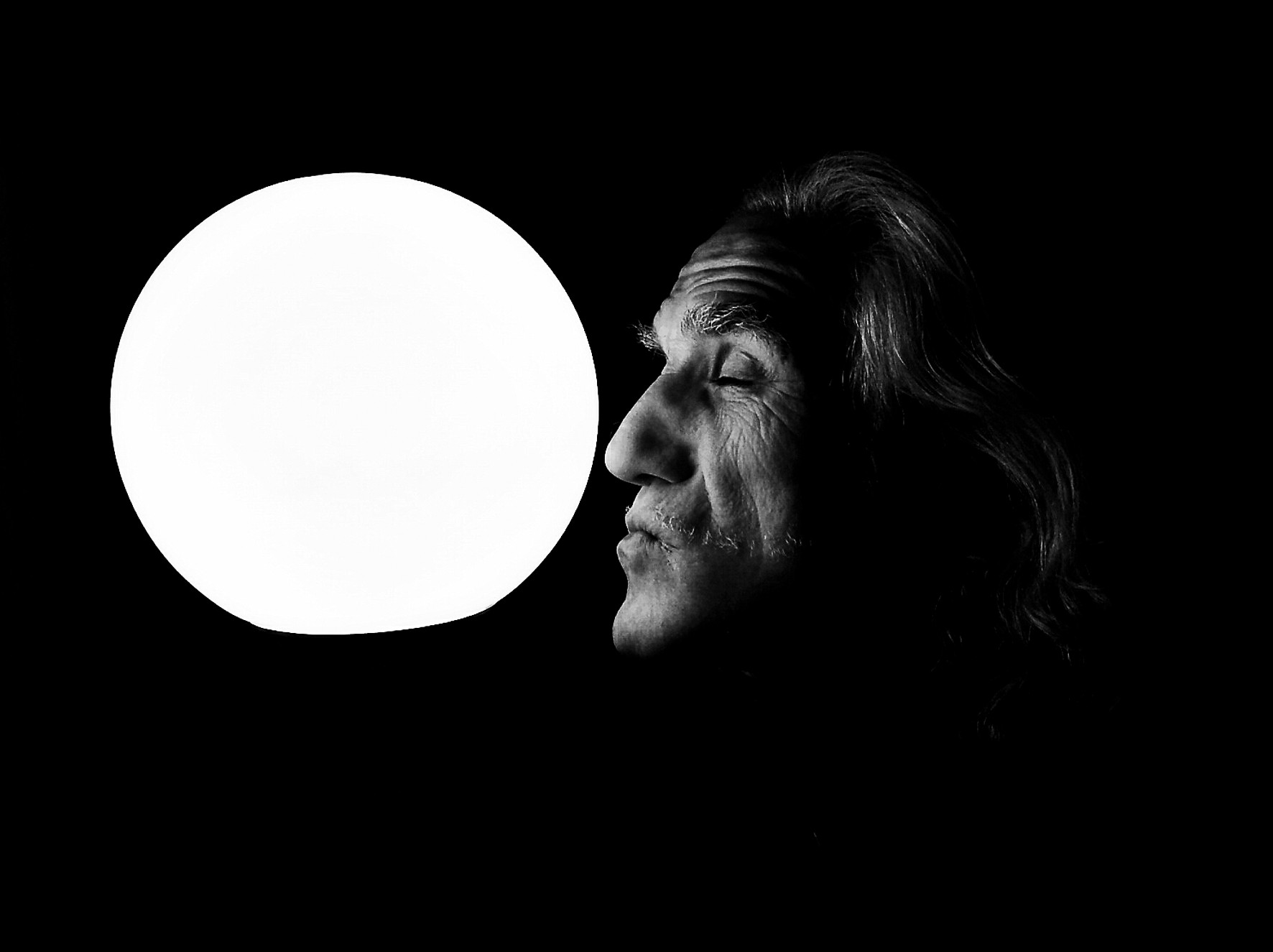 Portrait of an older man with long hair, in profile and eyes closed, facing a white circle on a black background.