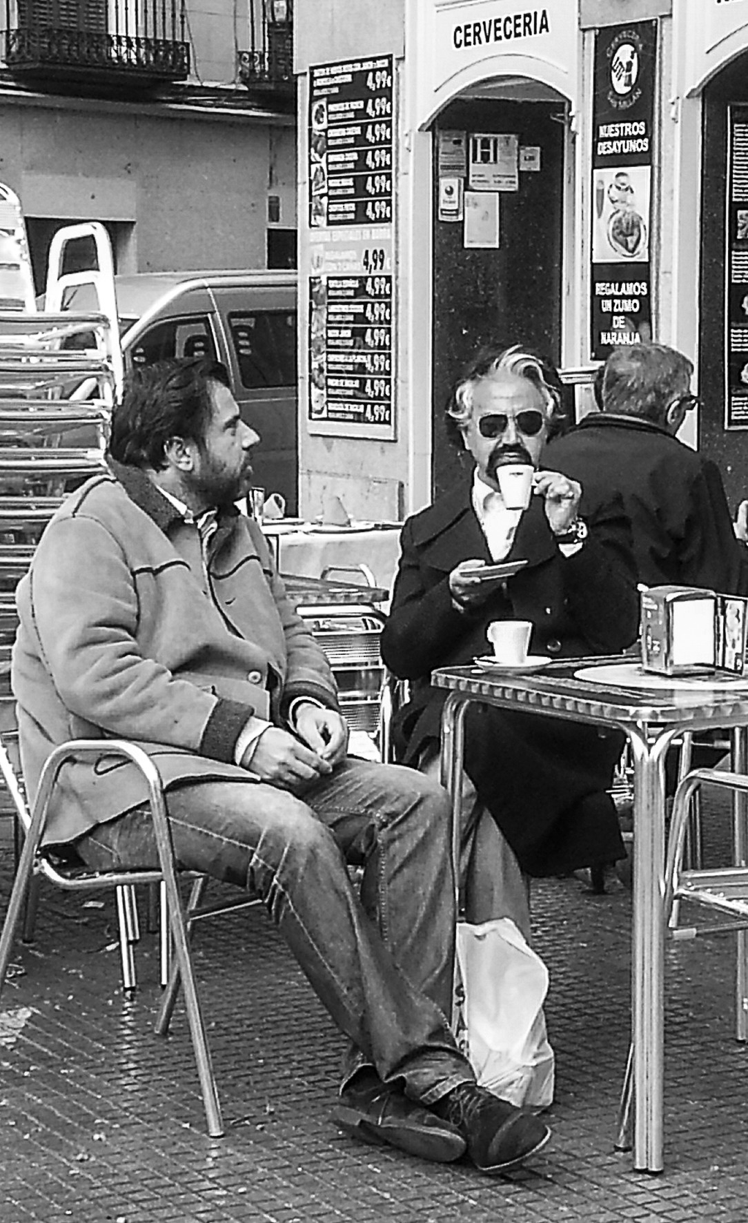 Two men seated at a café terrace table with coffee, in front of a bar.