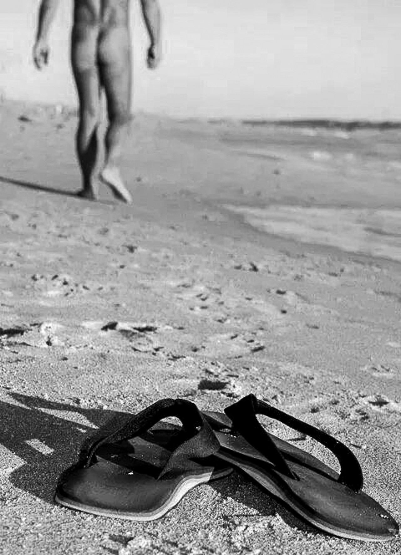 A nude man seen from behind walking along a beach shoreline; flip-flops in the foreground.