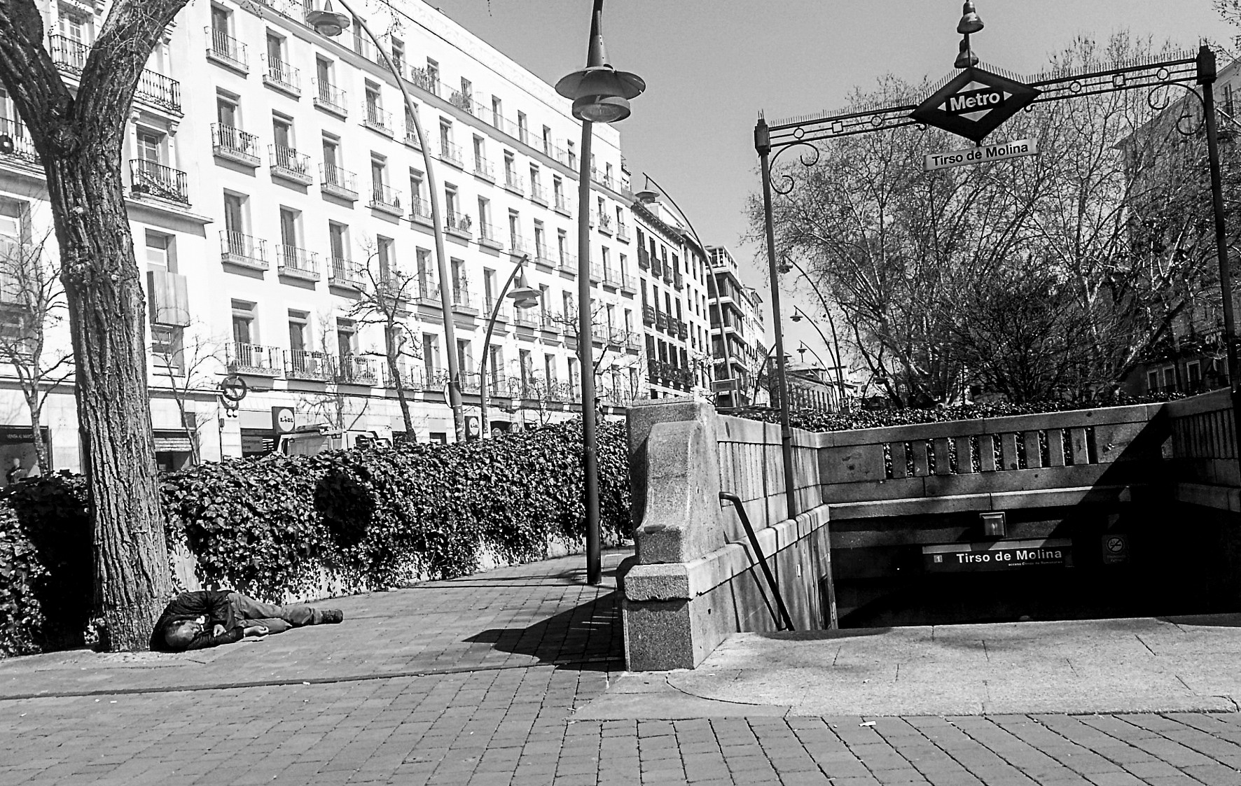 An empty pavement beside the Tirso de Molina metro entrance, with trees and apartment buildings.