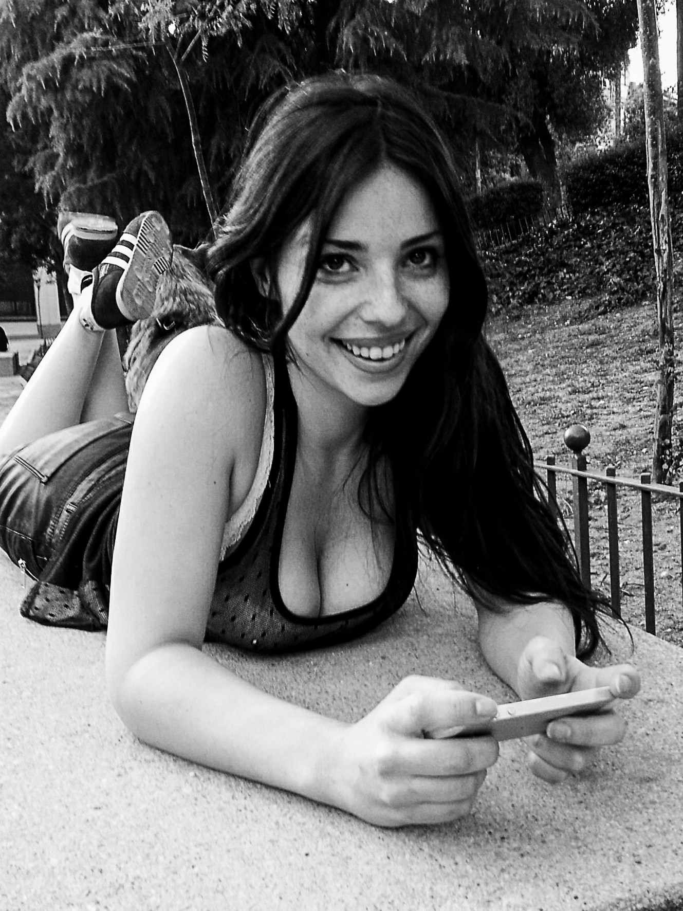 A young woman smiles at the camera, lying face-down on a wall, phone in hands.