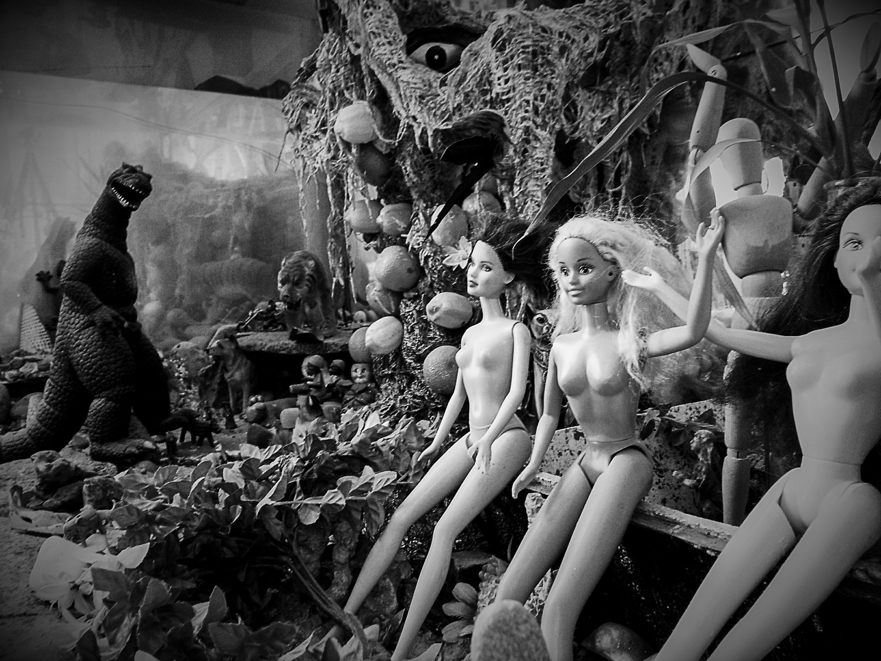 A shop display of nude dolls and a Godzilla figure among plants and plastic fruit.