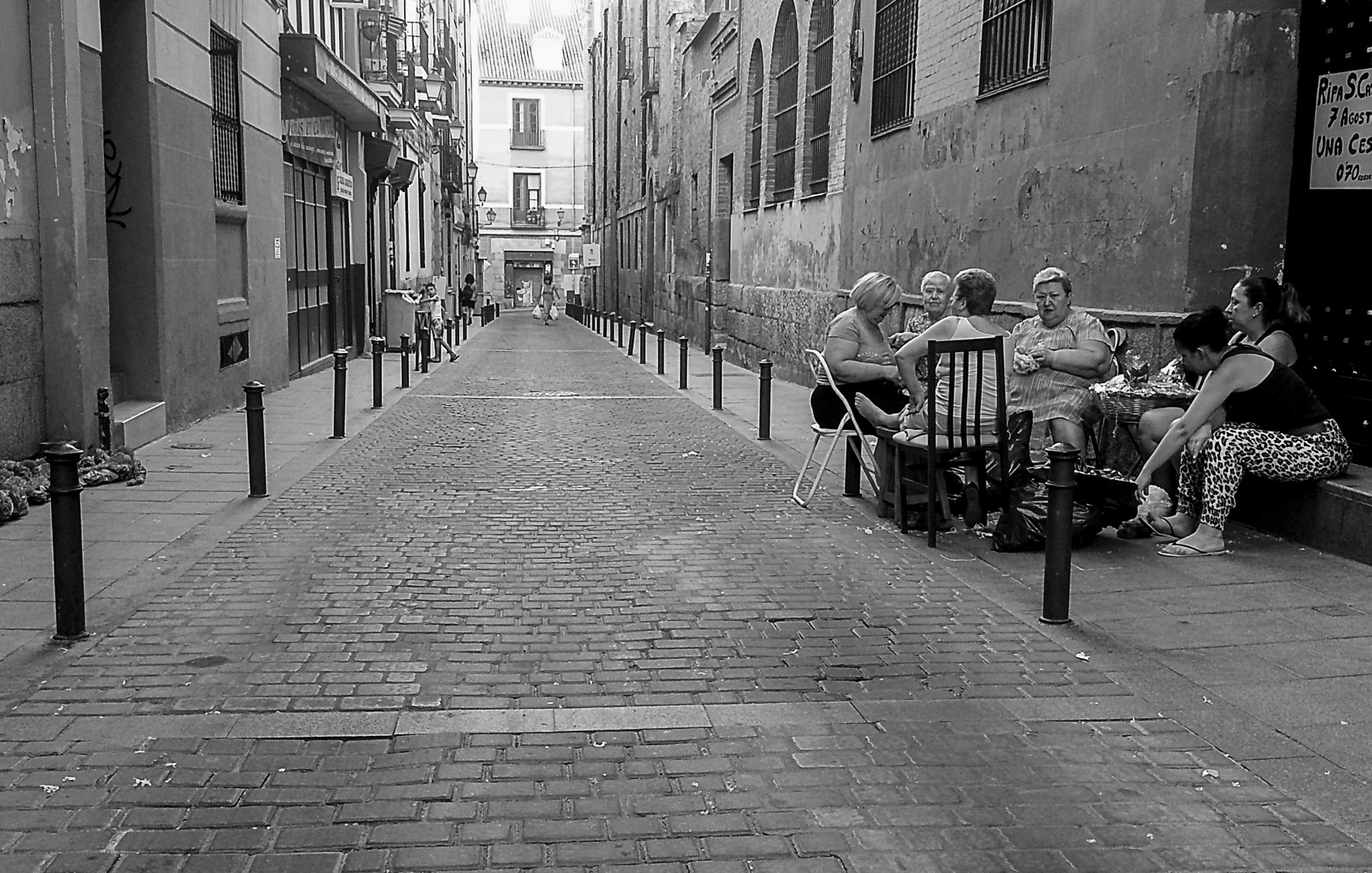 A narrow cobbled street with bollards and, on the right, a group of women seated on chairs at a doorway.