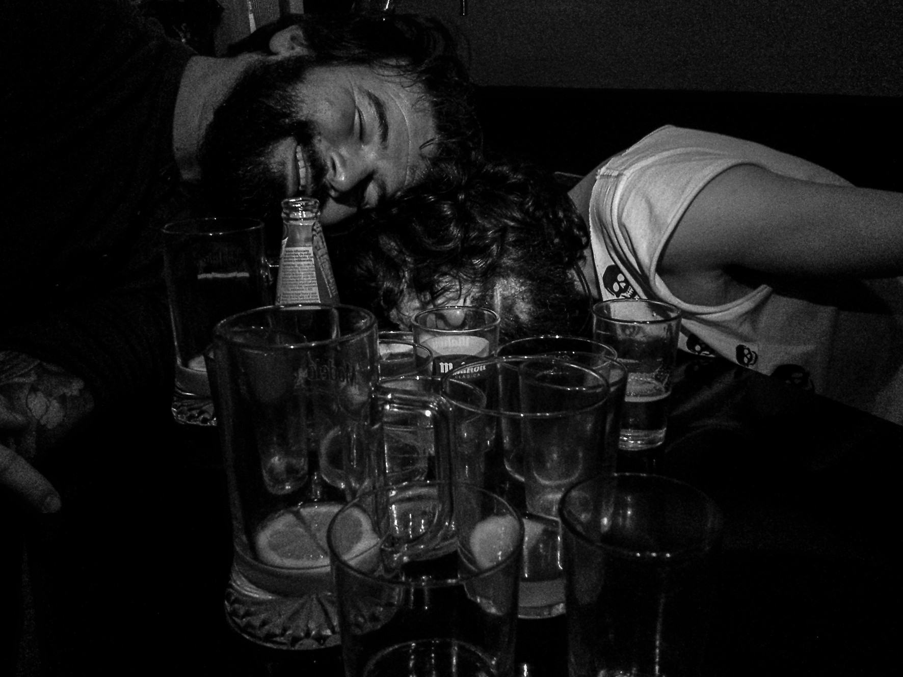 A man asleep with his head on a table, among empty glasses and bottles.