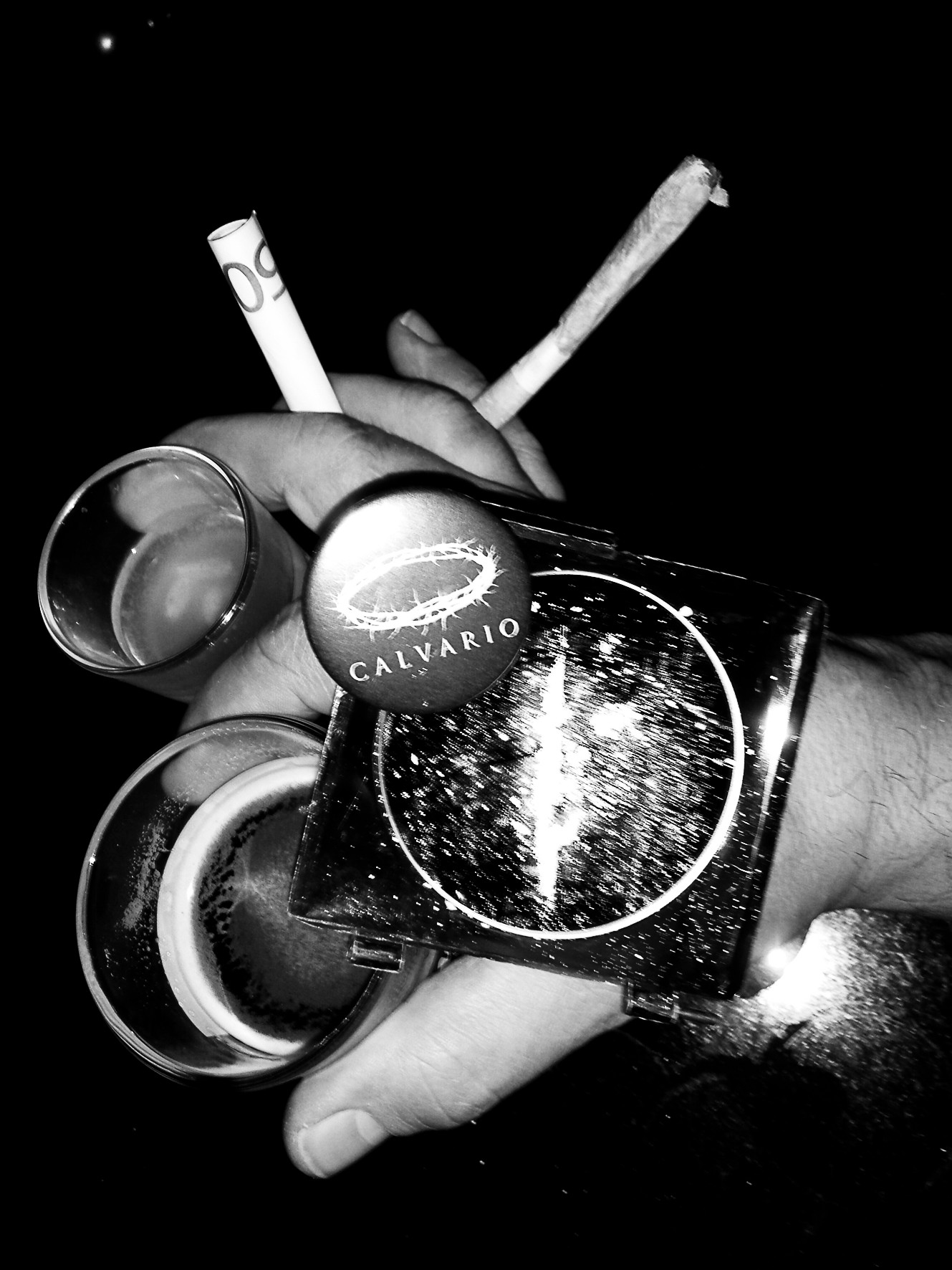 A hand with two rings holding a cigarette and a joint, wearing a wide cuff marked "Calvario".