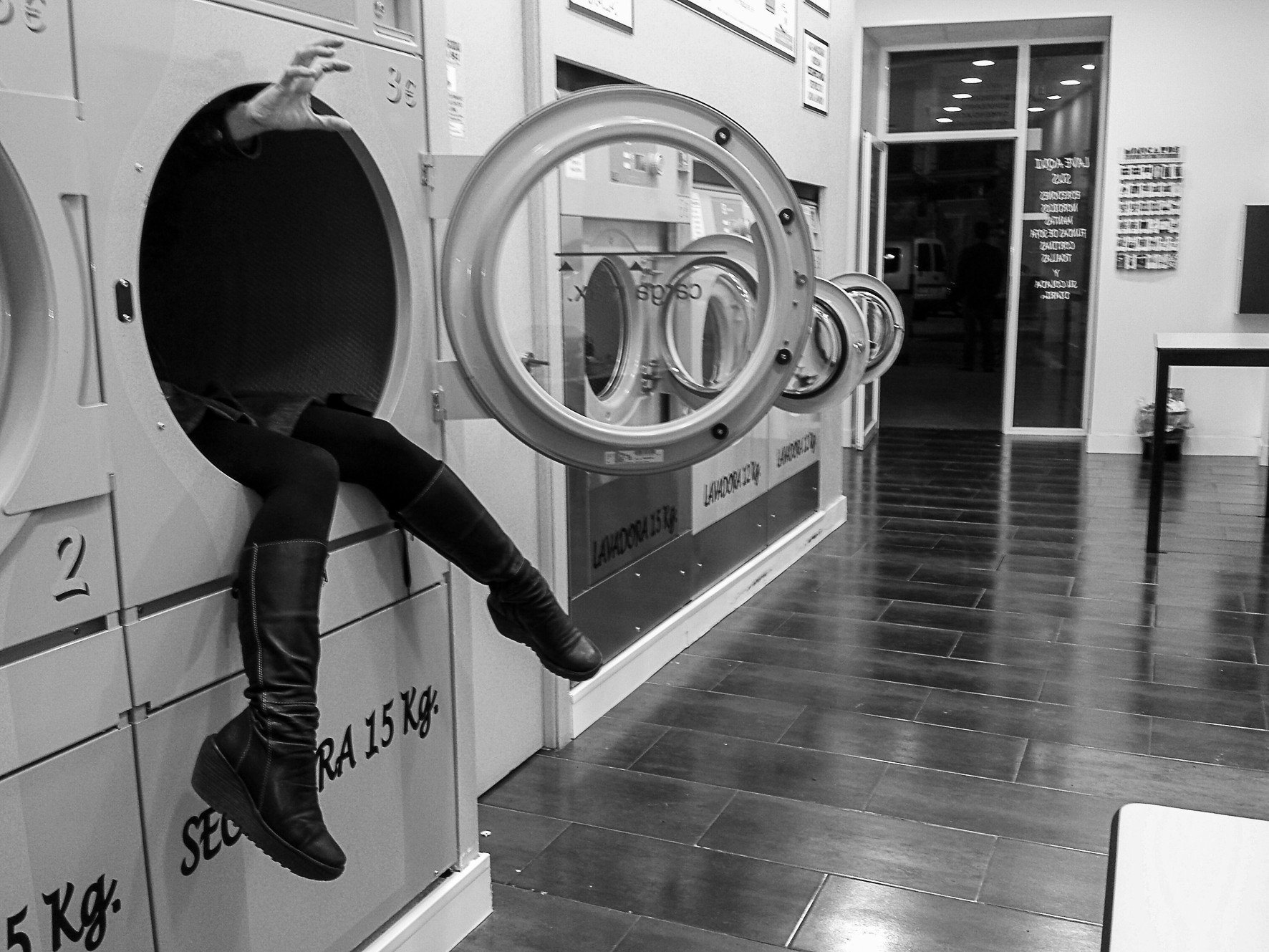 A woman climbs out of an industrial washing machine in a laundromat, her boots hanging out.