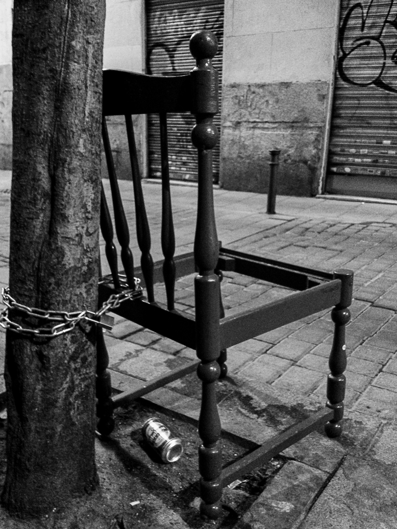 A wooden chair chained to a tree on the pavement, a can on the ground.