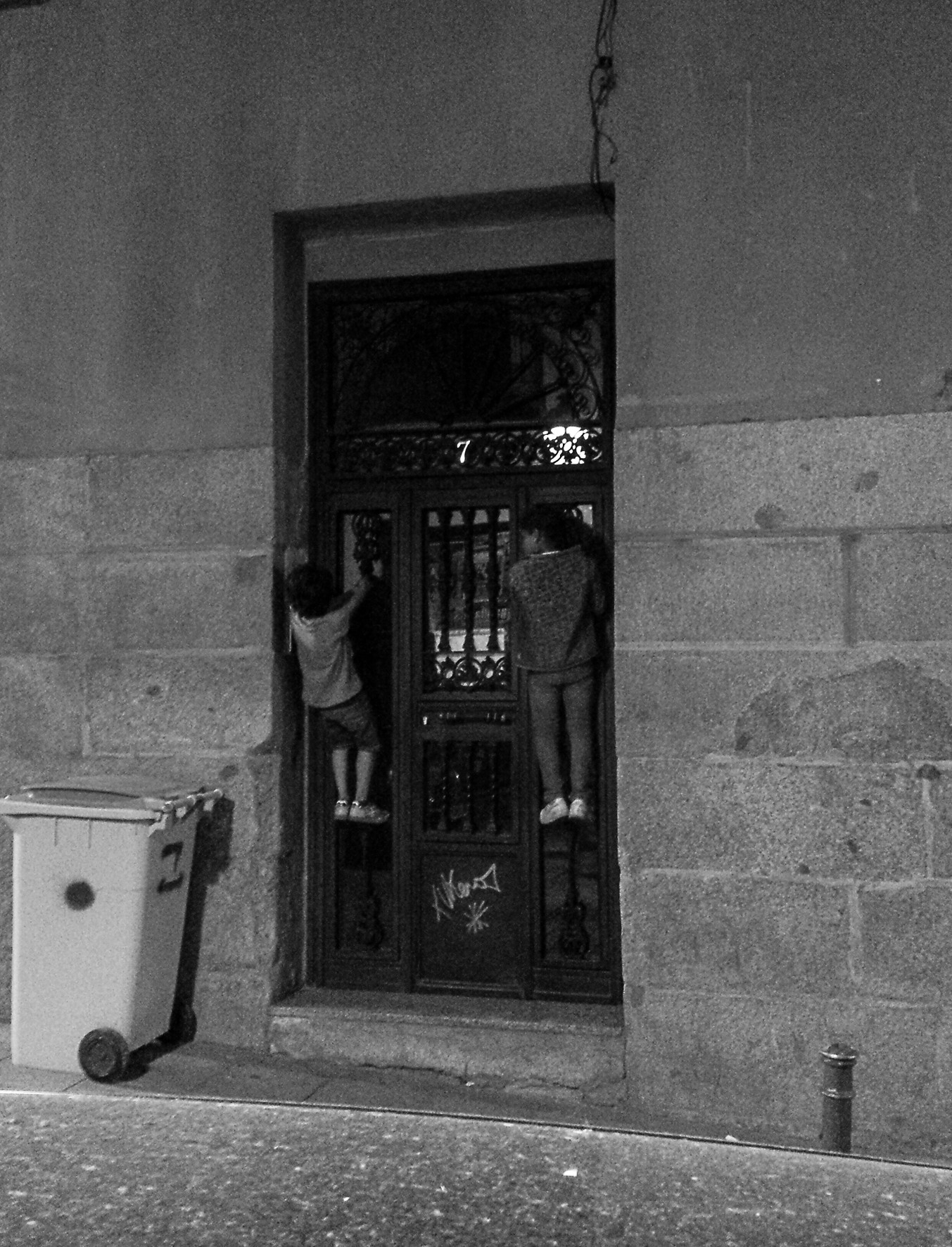 Two children climb the iron grille of a door in a stone wall, beside a bin.