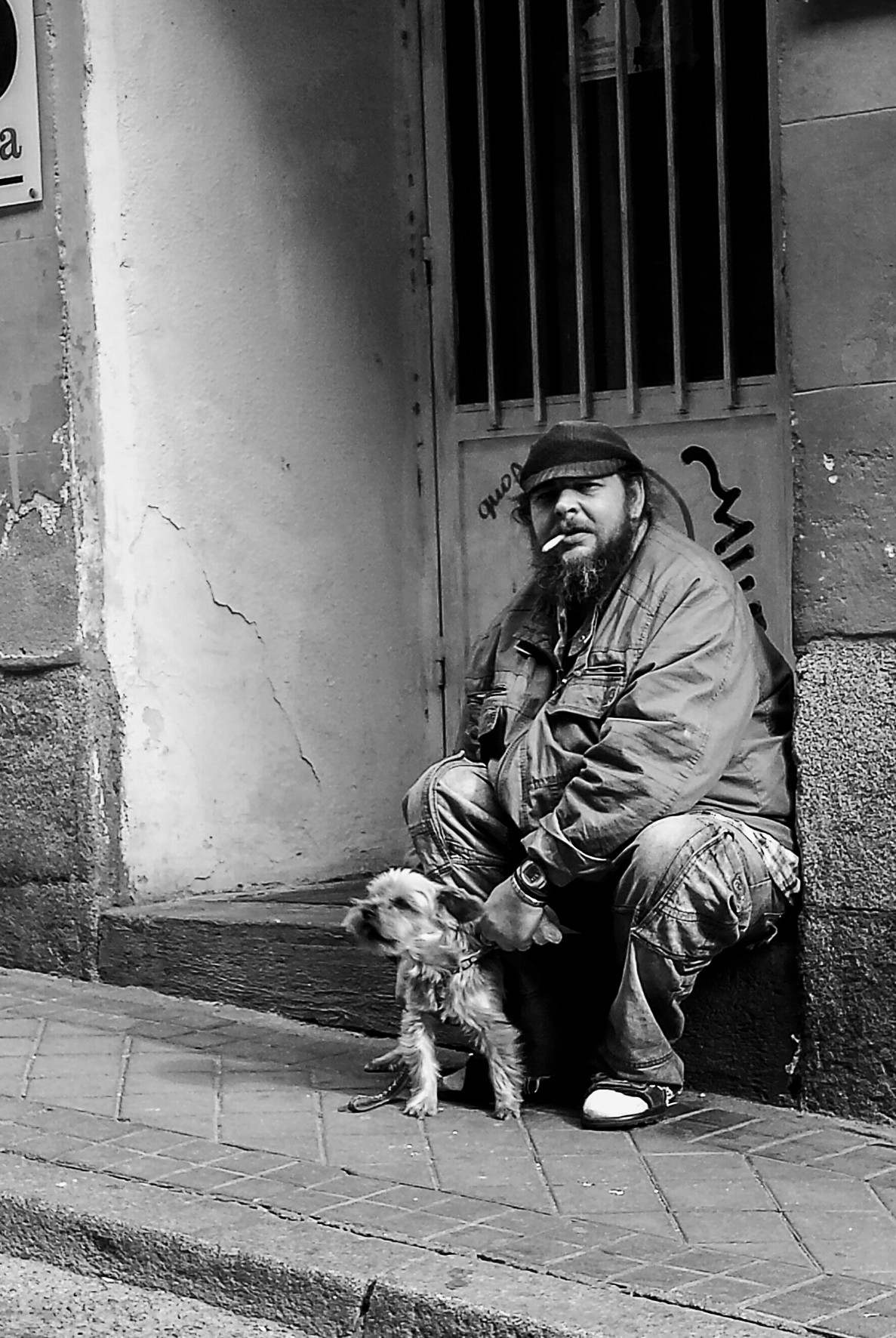 A man in a cap seated in a doorway with a small dog, smoking.