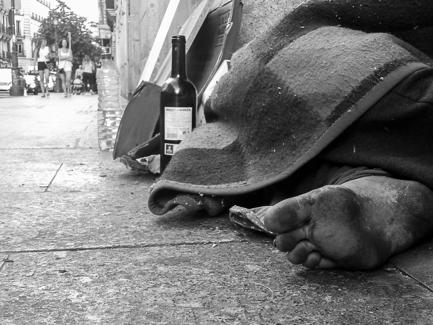 A bare foot sticks out from a blanket on the pavement, beside a wine bottle.