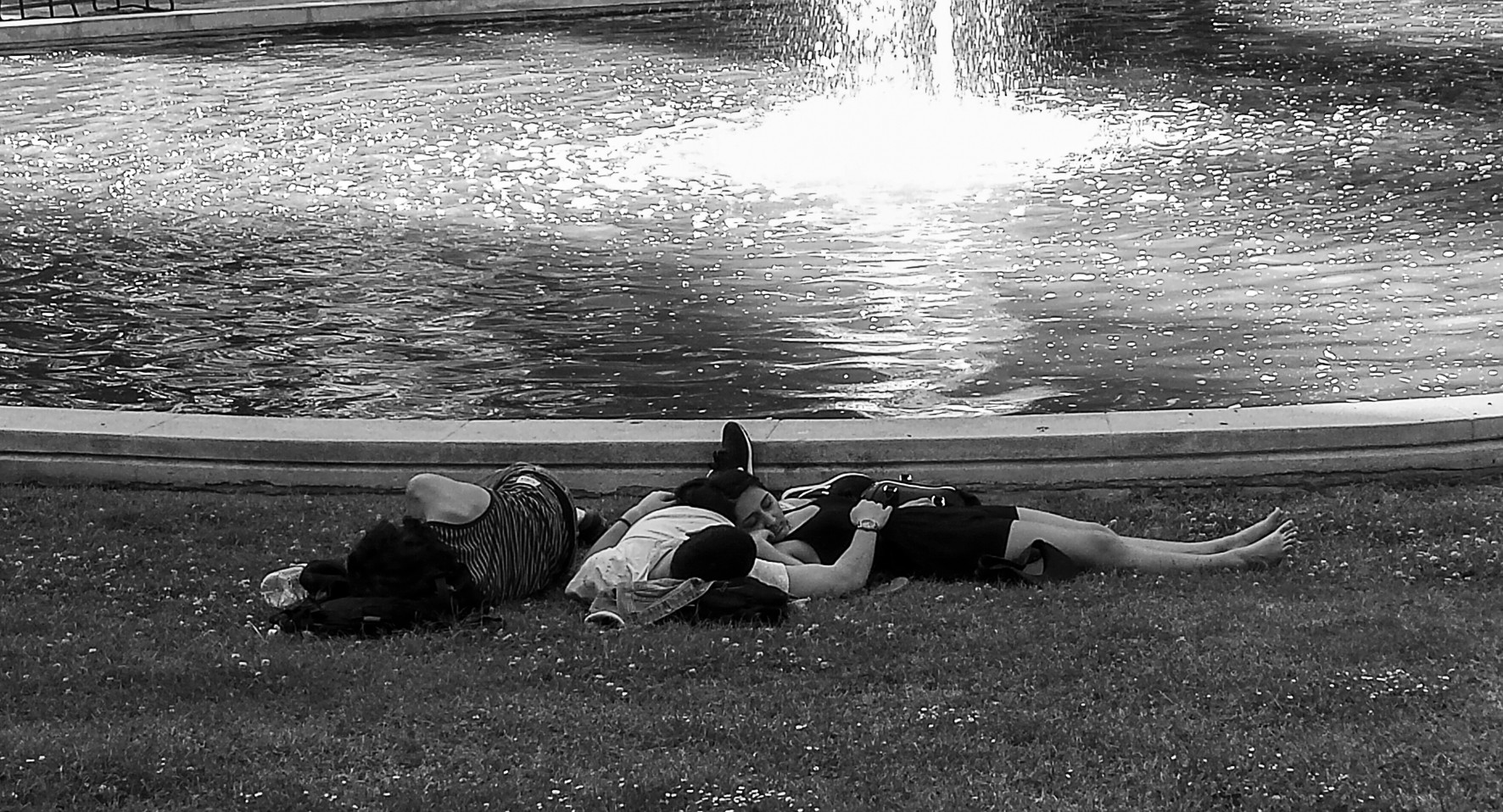 A group of people asleep on the grass beside the edge of a pond with a fountain.