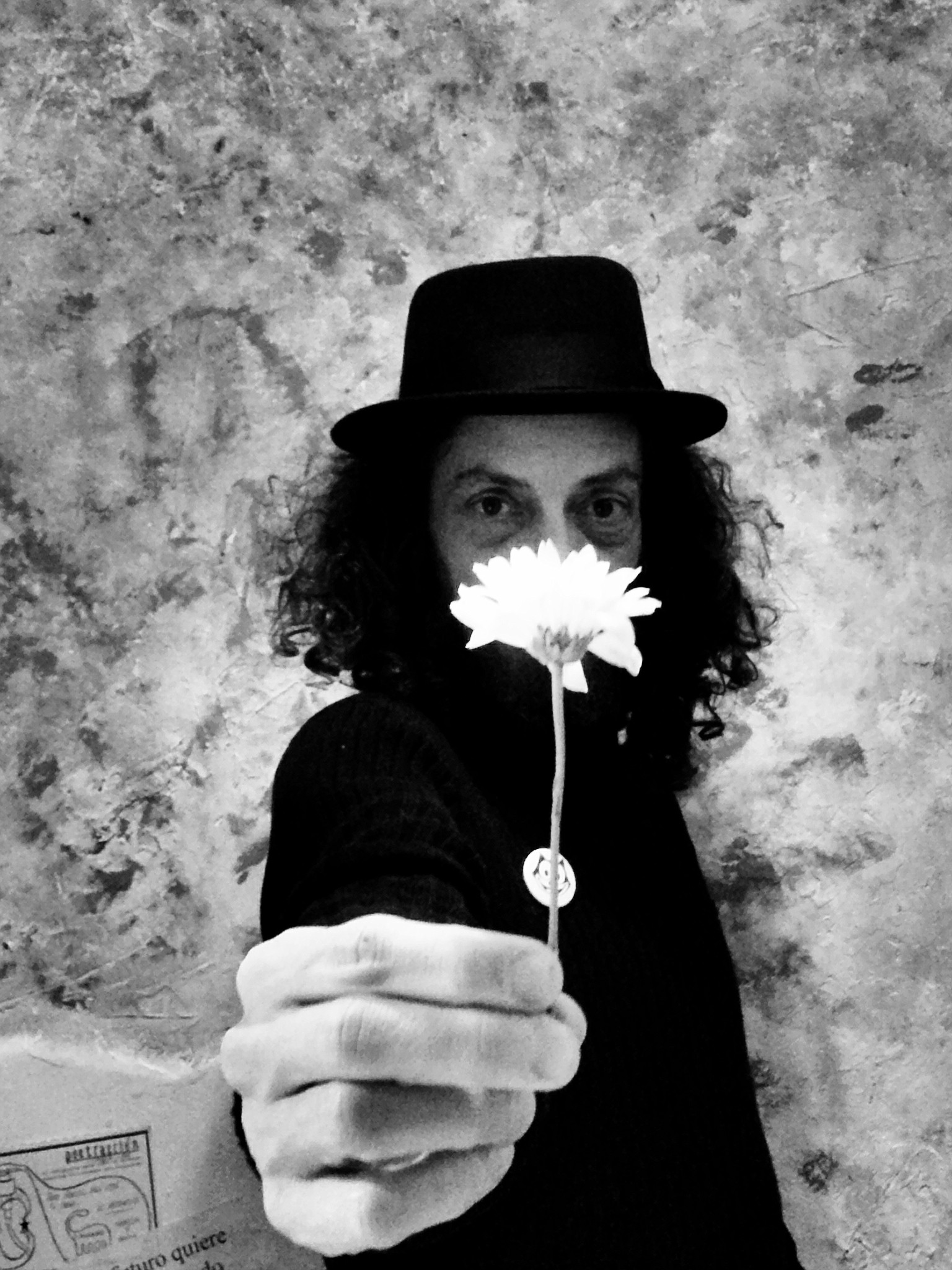 A man in a hat with curly hair covering his face with a flower held before him.