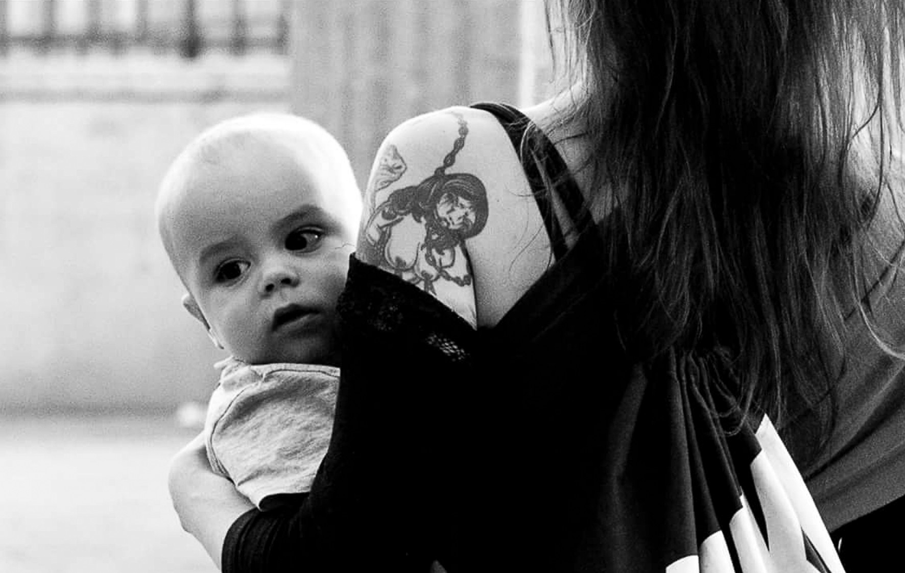 A baby looks at the camera from the shoulder of a tattooed woman who faces away.