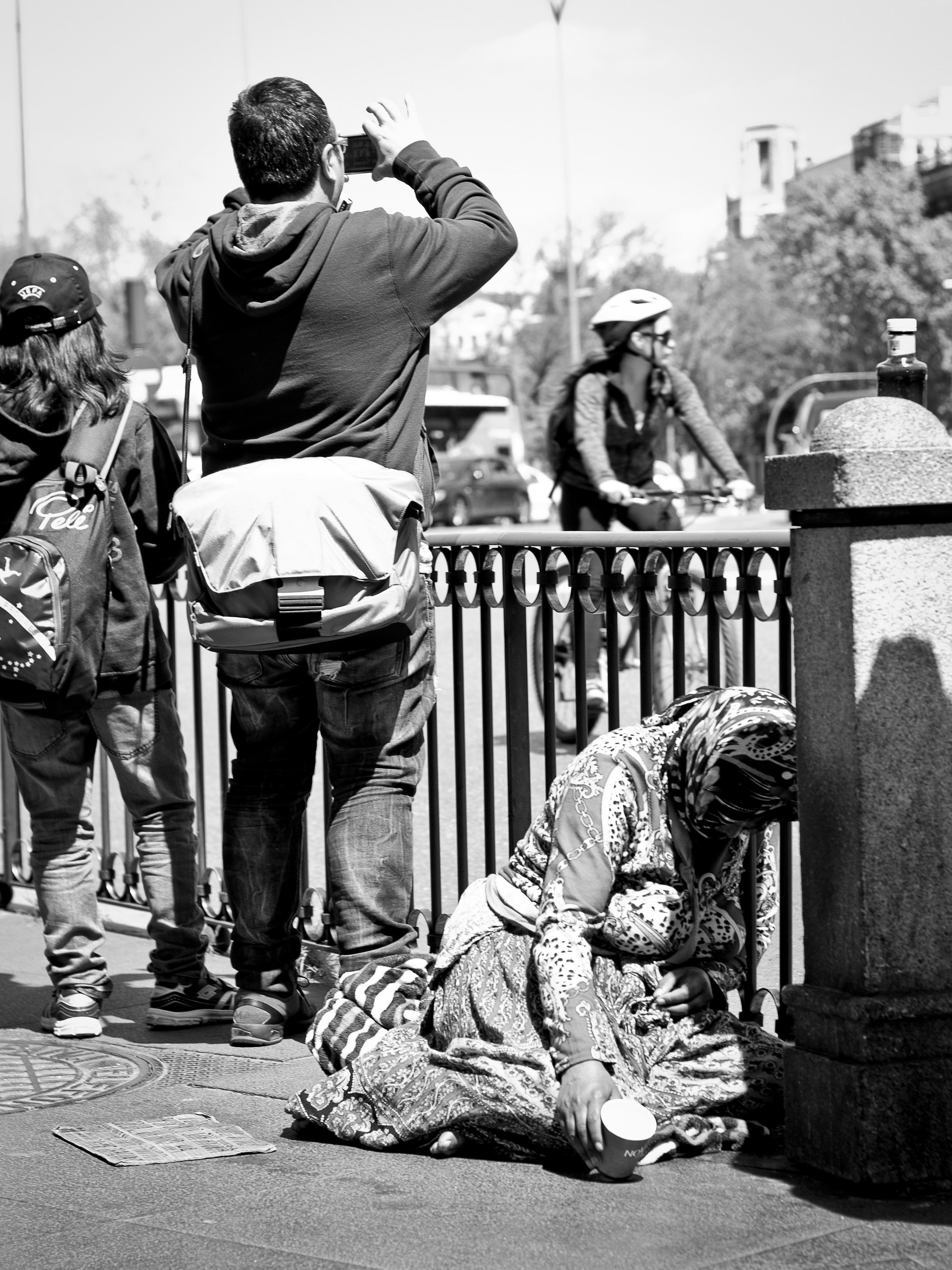 A group of tourists photographs beside a railing while a woman kneels on the ground begging.