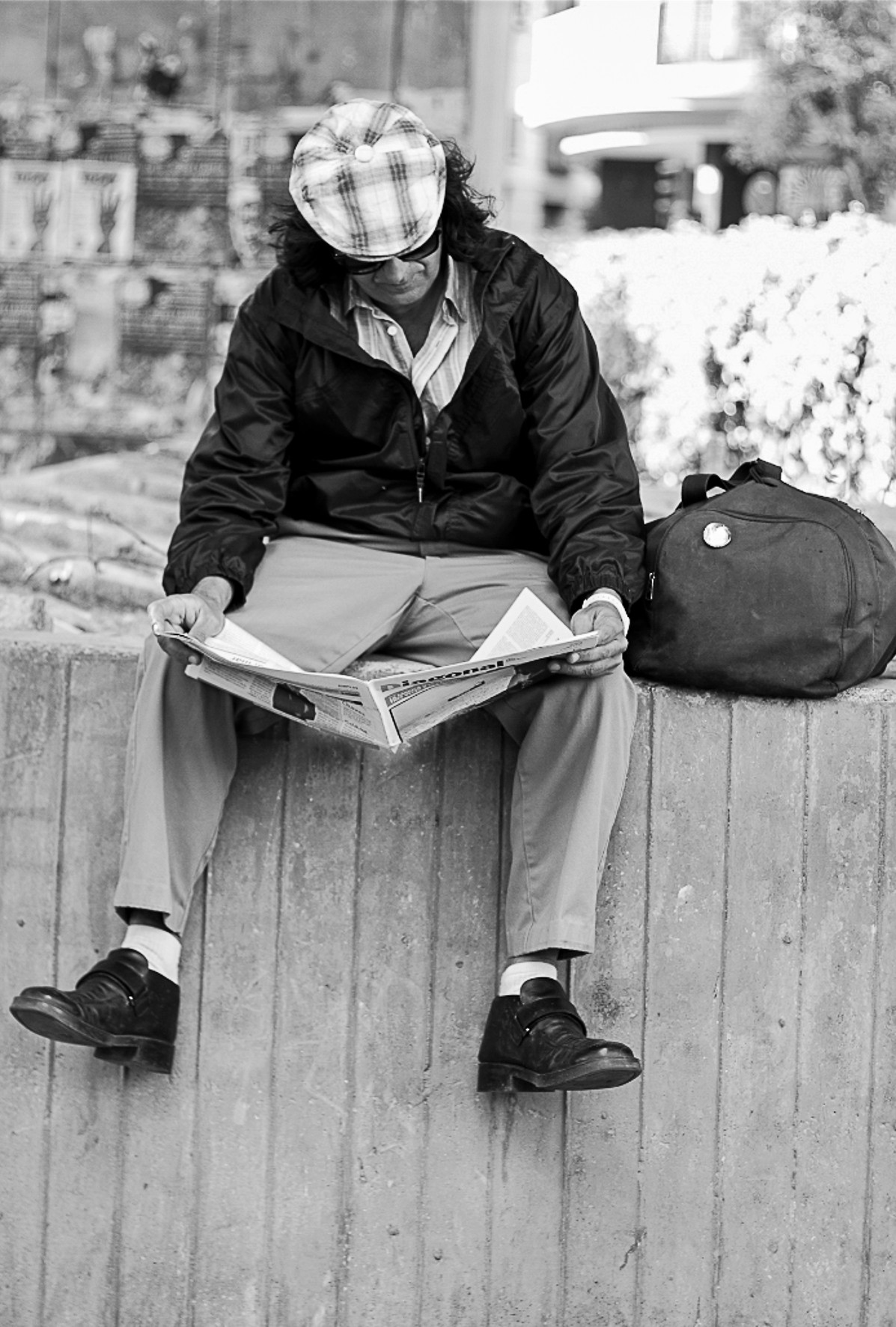 A man in a checked cap reads a newspaper seated on a wooden wall, a backpack beside him.