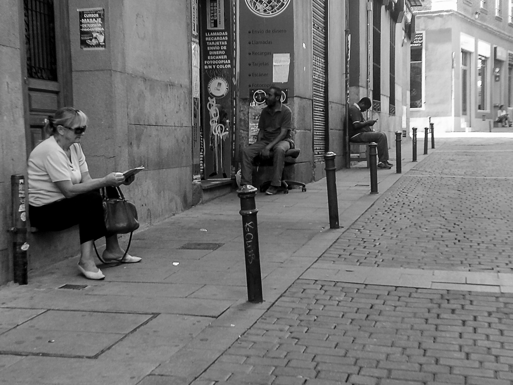 An older woman seated with a handbag on a bench in a cobbled street, other people in the distance.