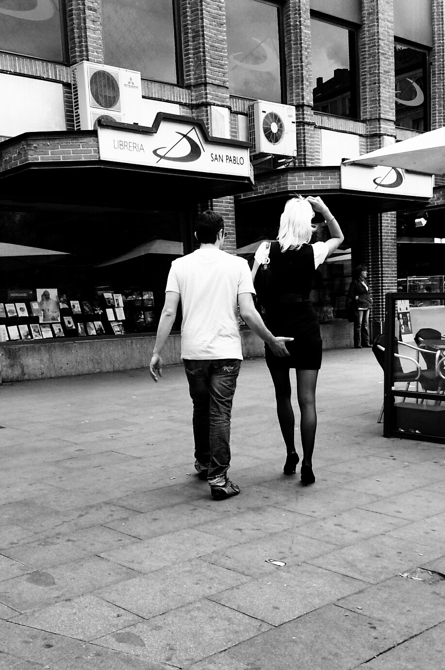 A couple seen from behind walking along a pavement beside a bookshop, she with black tights and blonde hair.