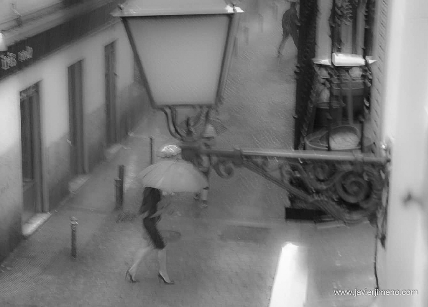 A woman in a skirt with a pale umbrella crosses a street in the rain, seen from above, beside a lamp.