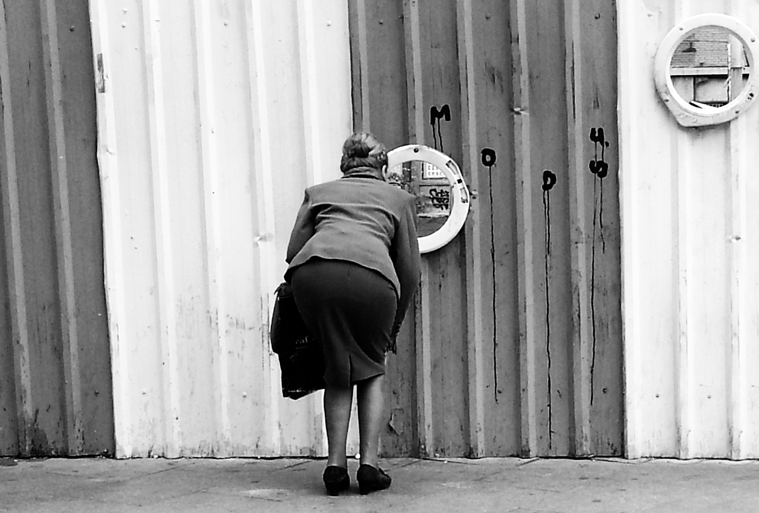 An older woman bends to look through a porthole in a graffitied metal fence; a second porthole at upper right.