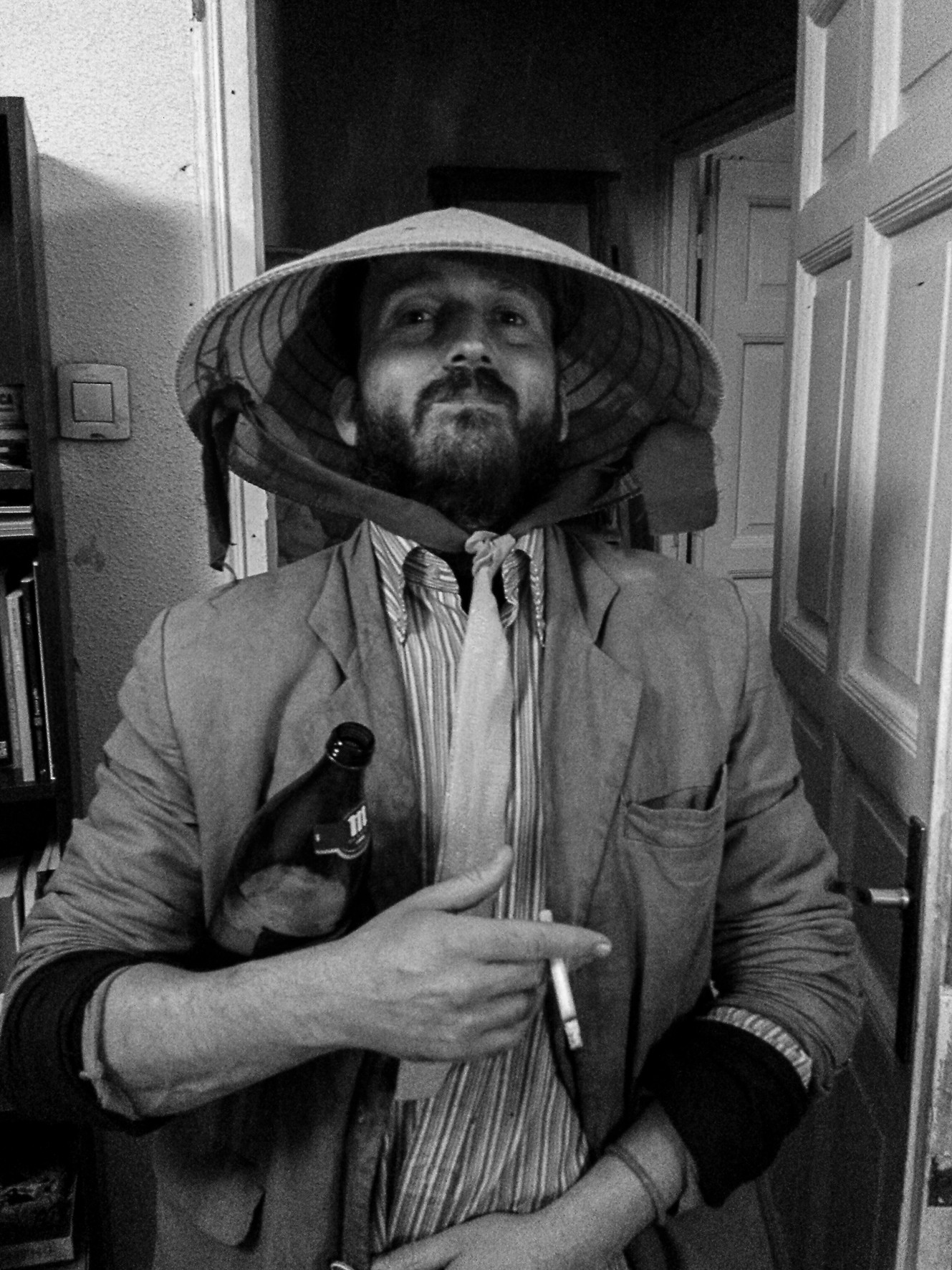 A man in a conical straw hat with a bottle, pointing at the camera with a cigarette, in a living room.