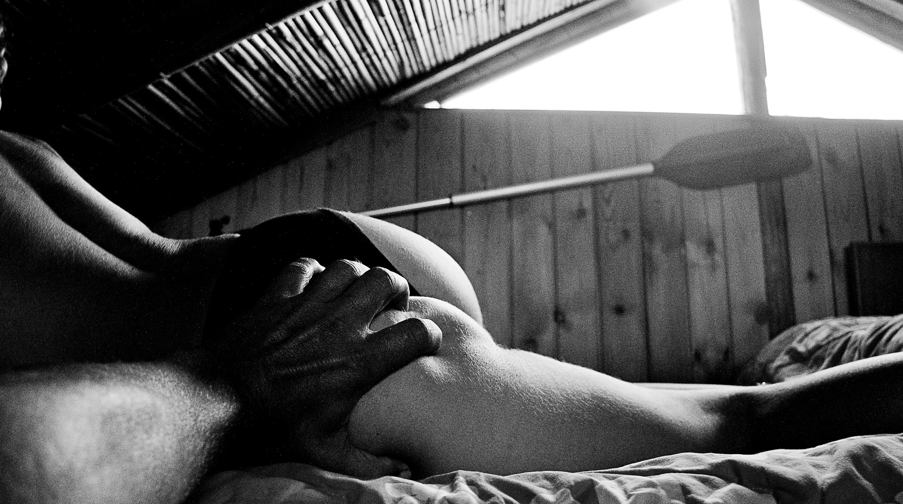 A nude body lying on a bed beneath a wooden ceiling, a hand on the hip and an oar behind.