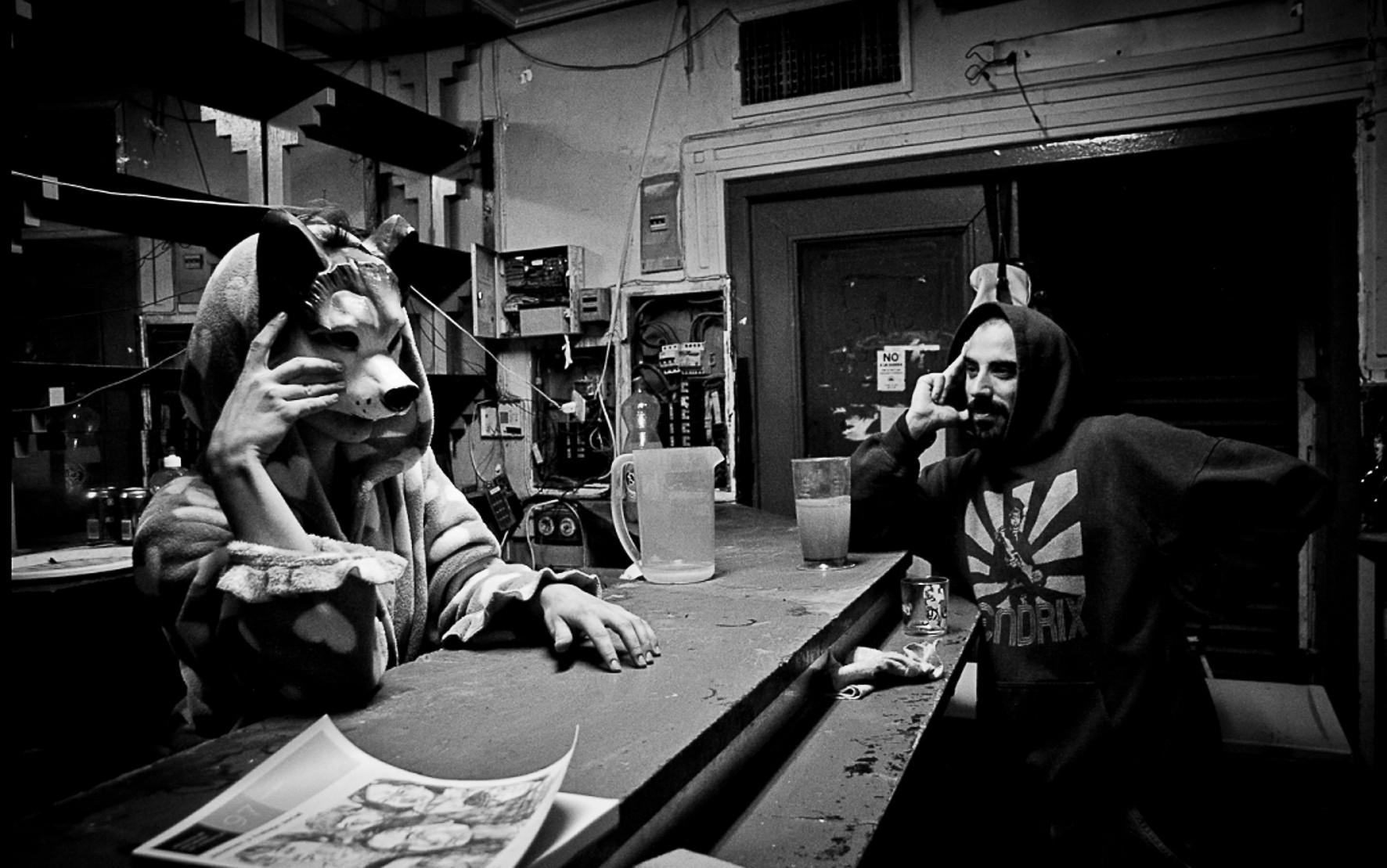 Two people, one in an animal mask and one in a hood, seated at a bar in a cluttered venue.